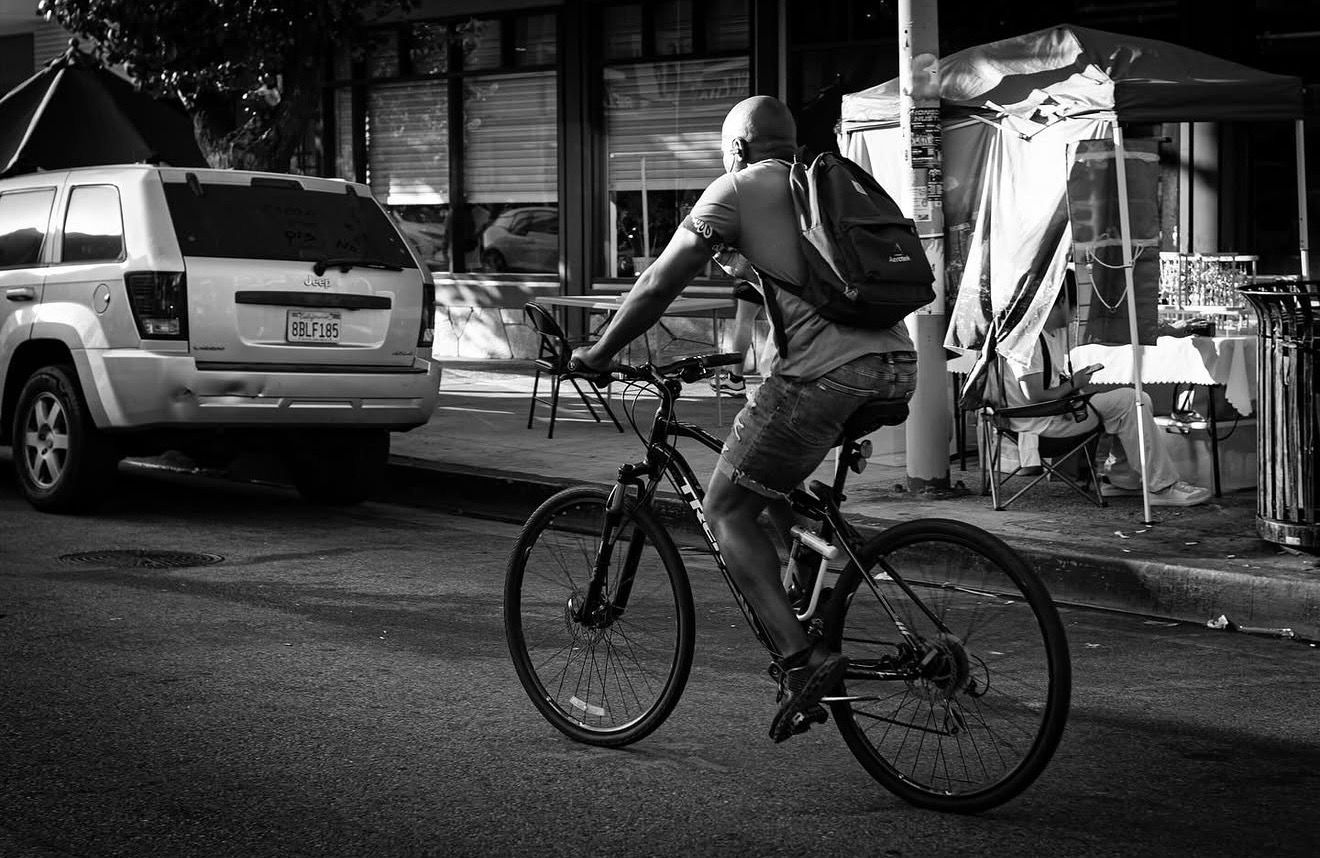 Black and white photo of a man riding a bicycle on a city street with a backpack and parked SUV nearby.