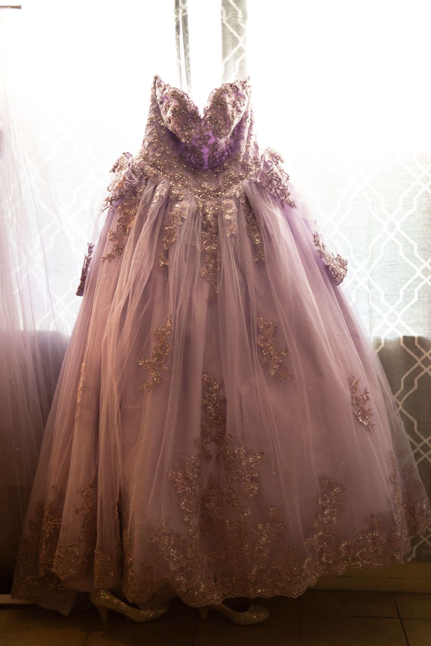 Elegant lavender ball gown with gold lace embroidery hanging by a sunlit window.