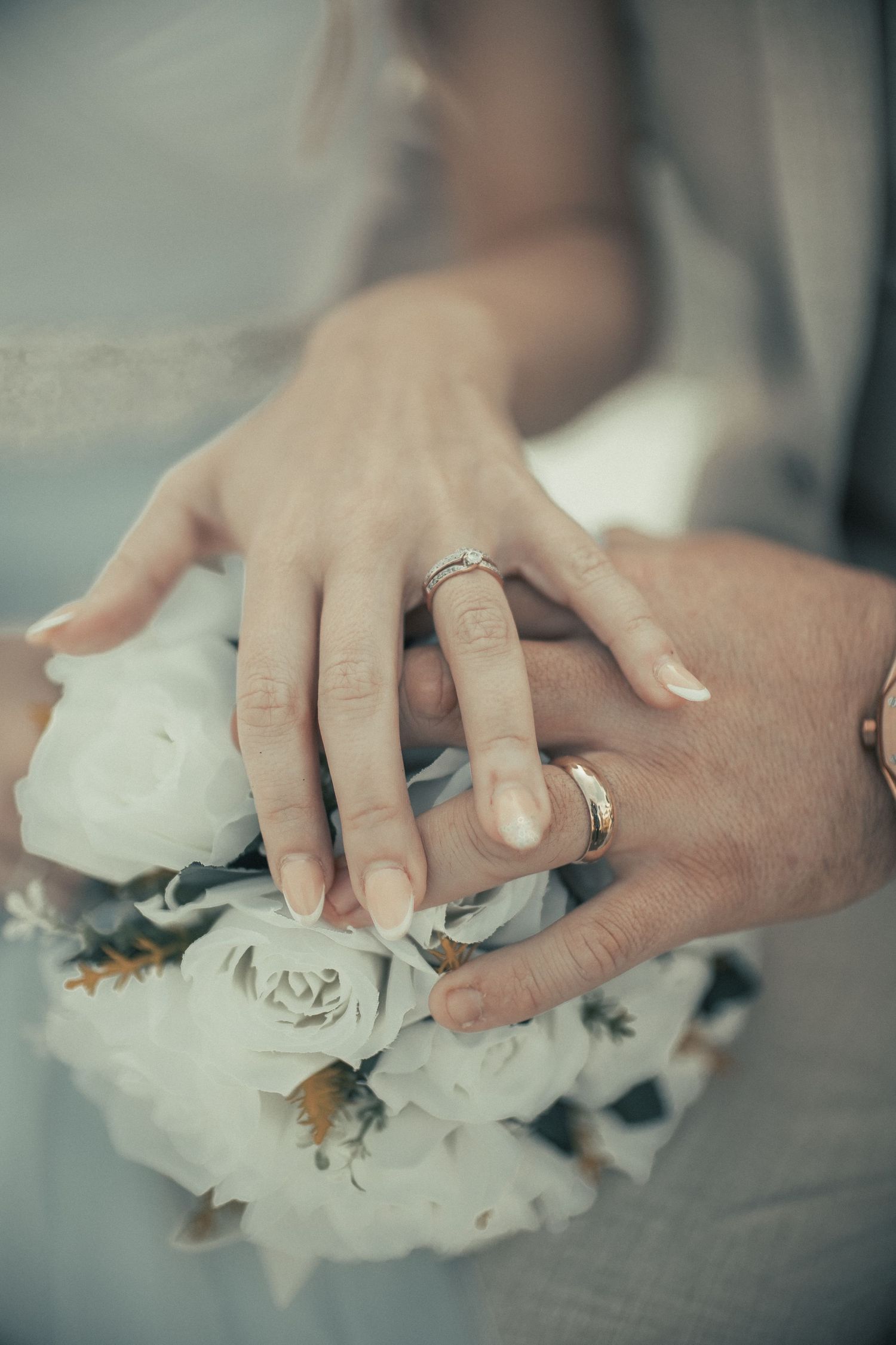 Close-up of bride and groom wedding rings on hands resting over white rose bouquet.