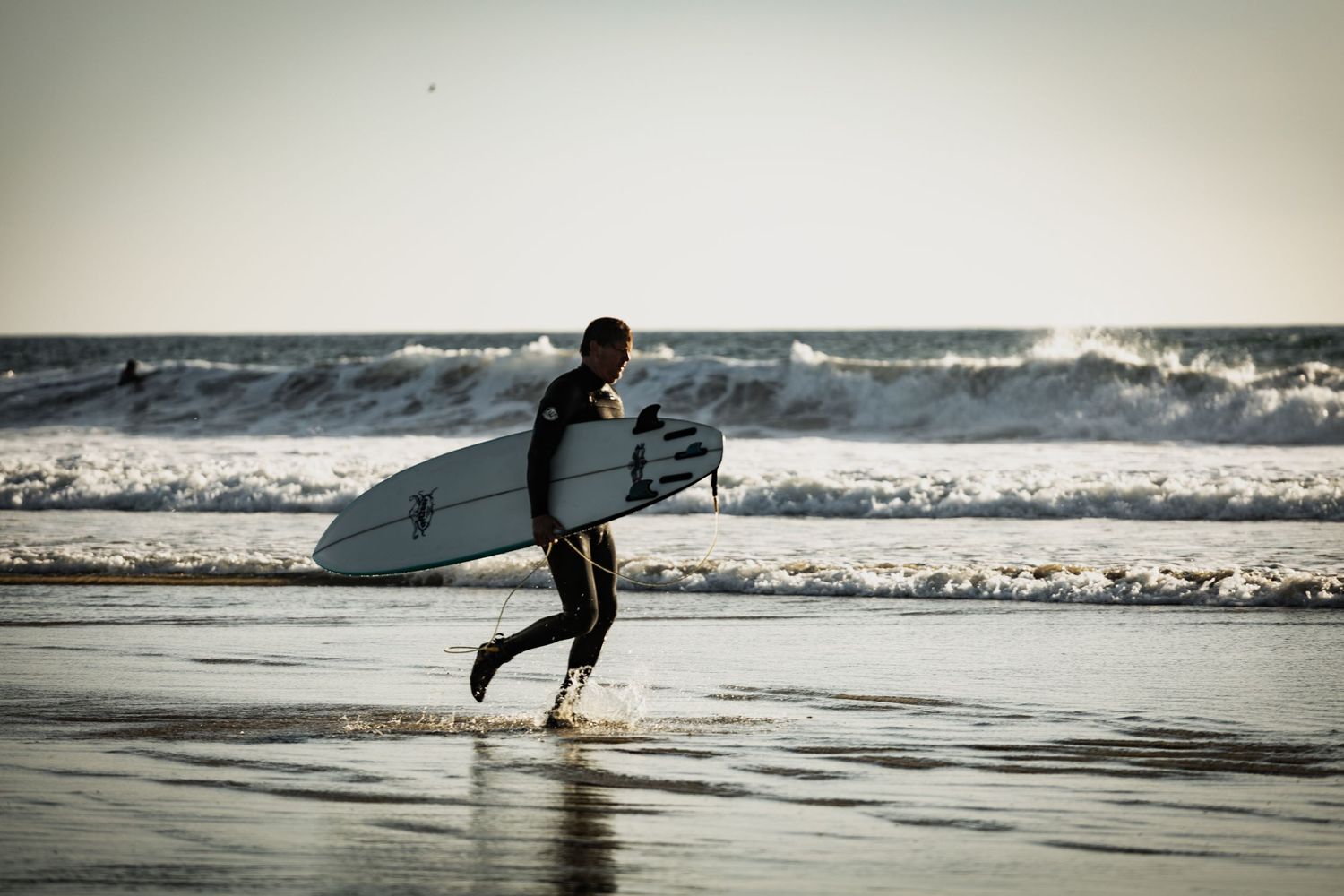 Surfer in a wetsuit carrying a white surfboard while walking along the shoreline toward ocean waves.