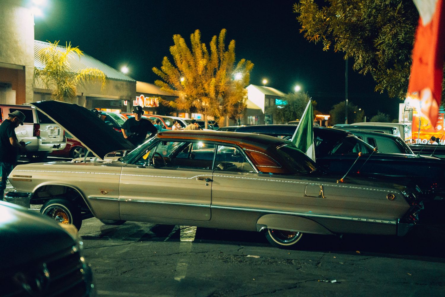Vintage silver lowrider car with open hood displayed in a parking lot during a nighttime car show.