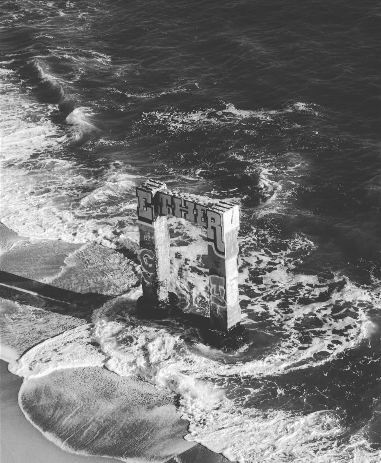 Aerial black and white photo of a lone concrete structure surrounded by crashing ocean waves.