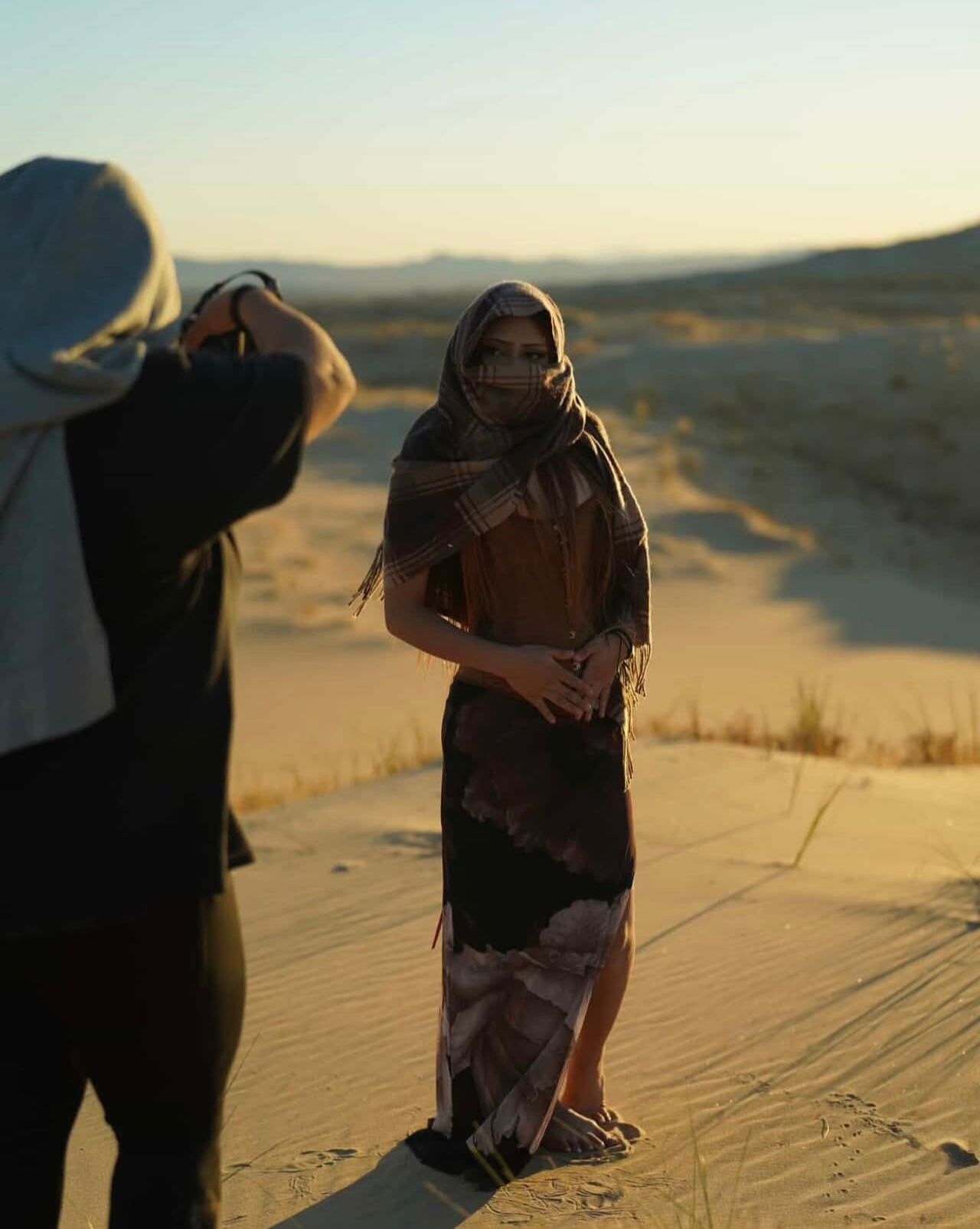 Photographer capturing woman in desert-inspired outfit with head scarf posing on sandy dunes at sunset.
