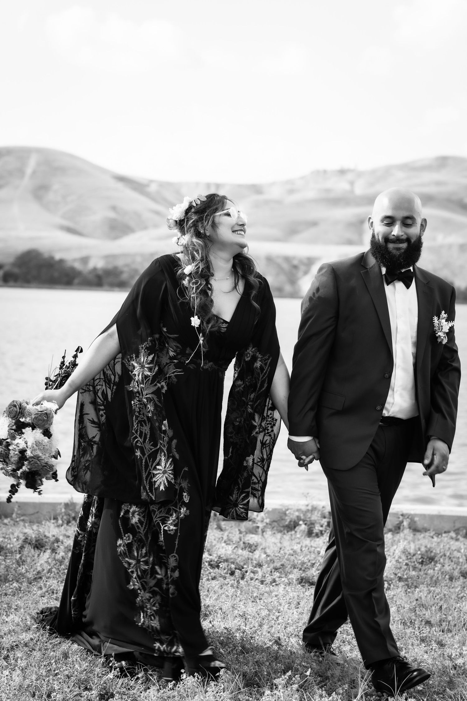 Black and white photo of a smiling bride in floral gown and groom in tuxedo walking hand in hand outdoors.