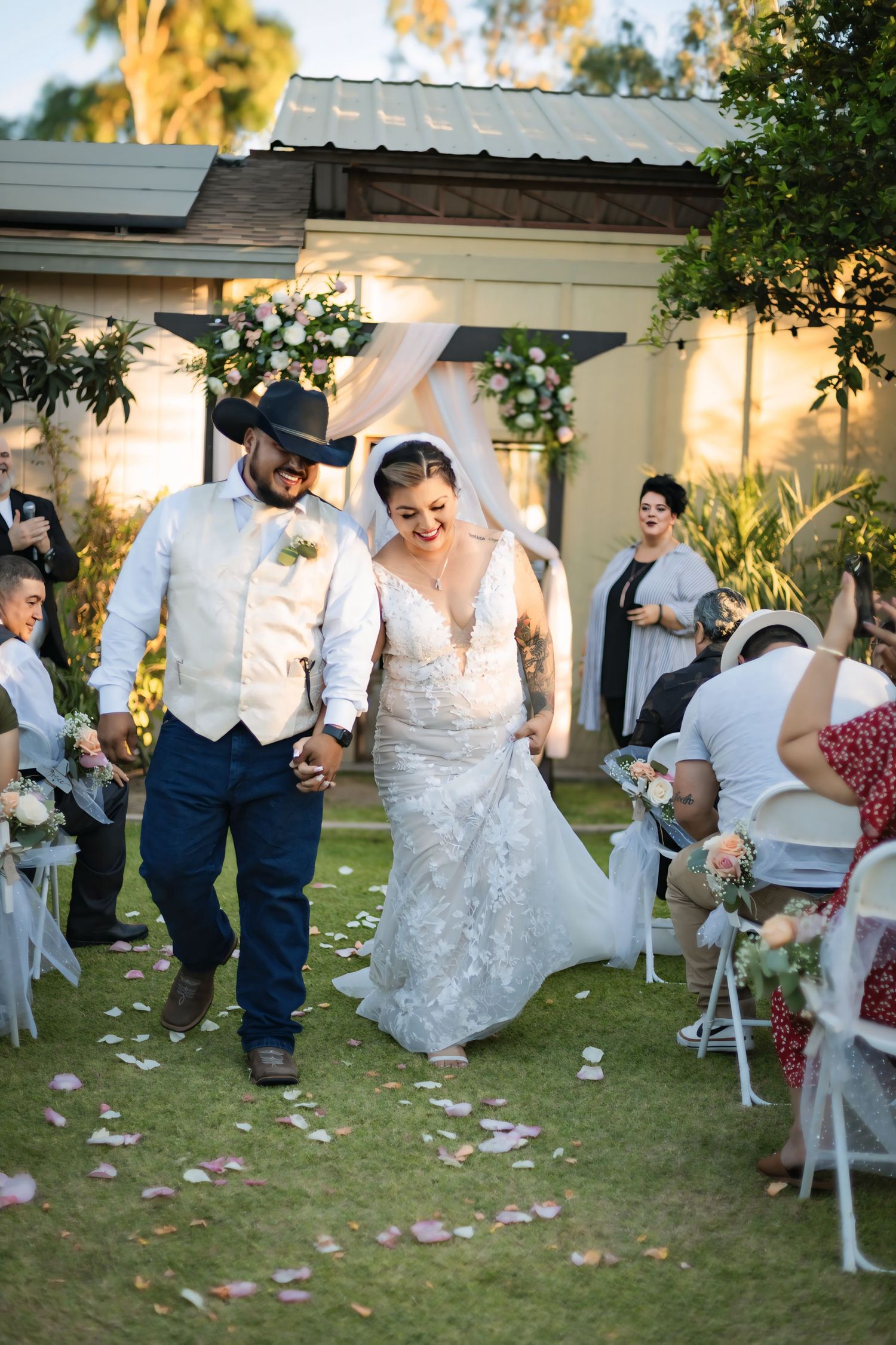 Bride and groom walk down the aisle after outdoor wedding ceremony, groom in cowboy hat and vest, bride in lace gown.