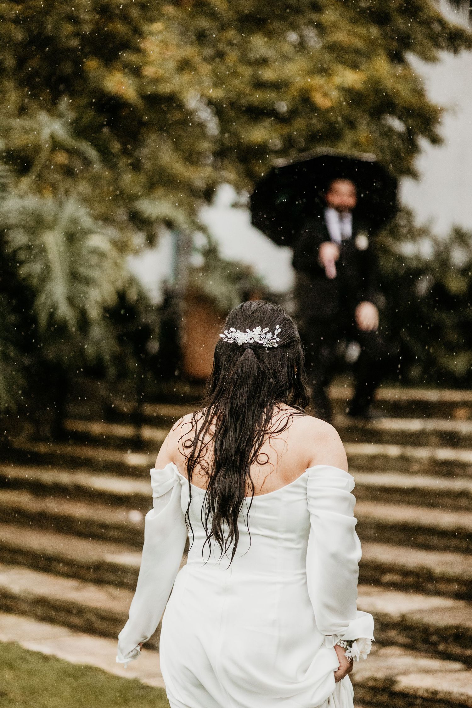Bride in white off-shoulder gown with floral hair accessory walks toward groom holding umbrella in rainy garden.