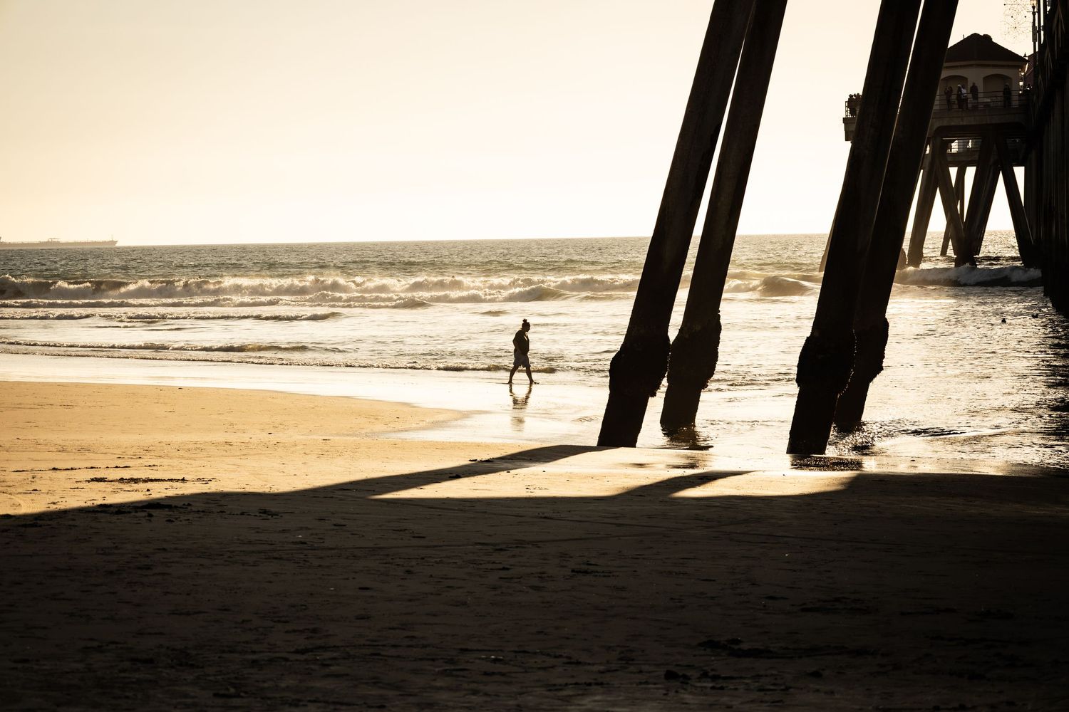 Silhouette of a person walking on a sunny beach near a pier with long shadows cast on the sand.