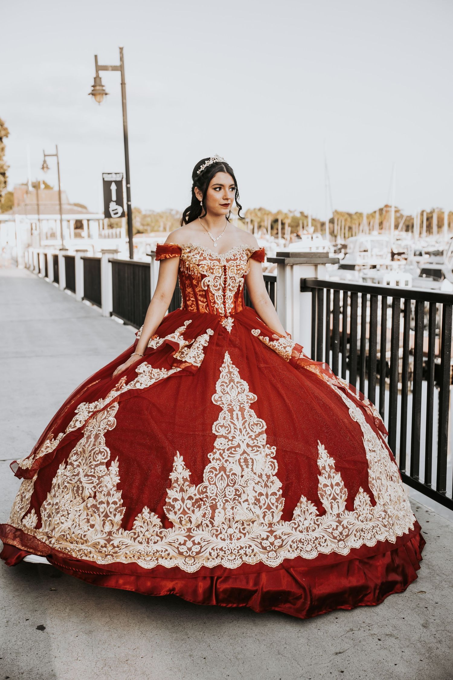 Young woman in stunning red and gold embroidered quinceañera ball gown posing on waterfront promenade.