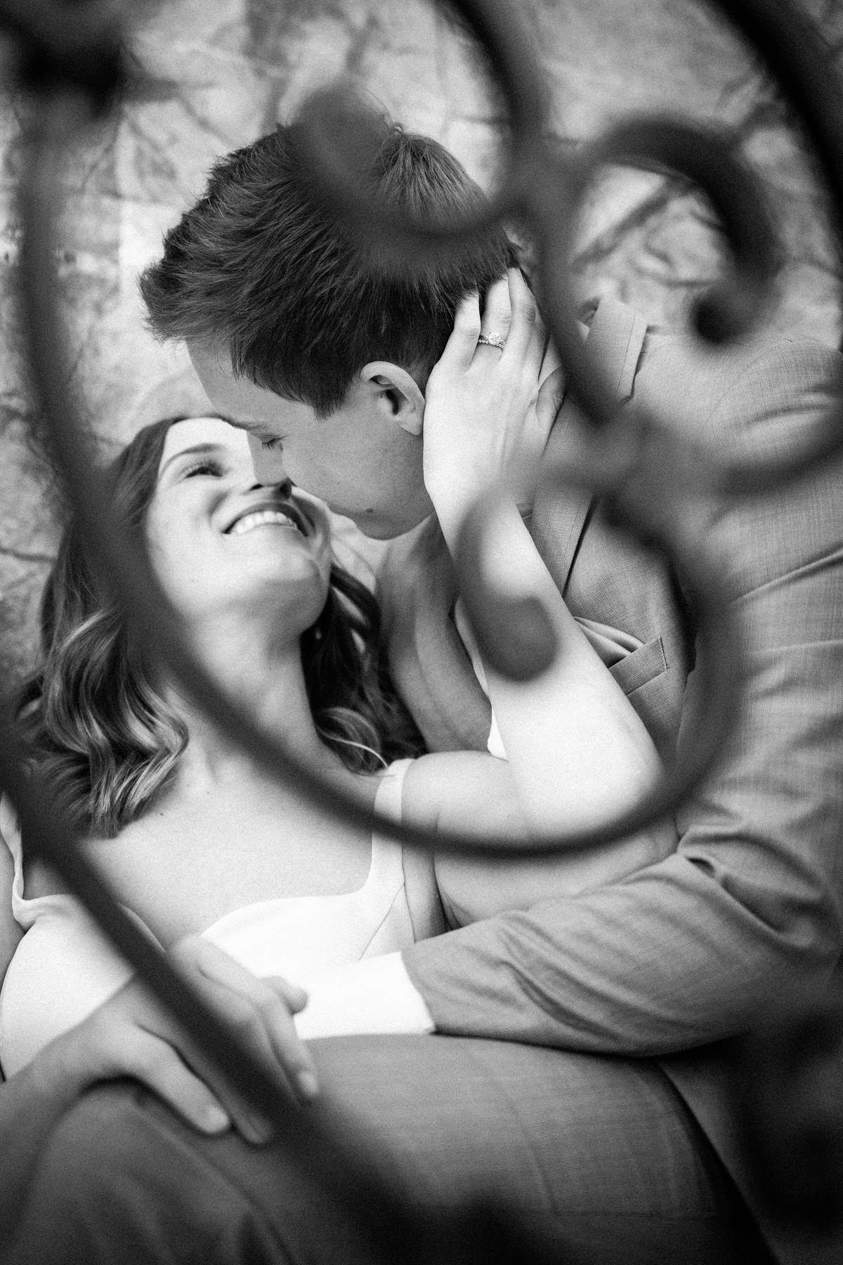 A smiling couple shares a tender moment framed by decorative iron scrollwork in black and white.