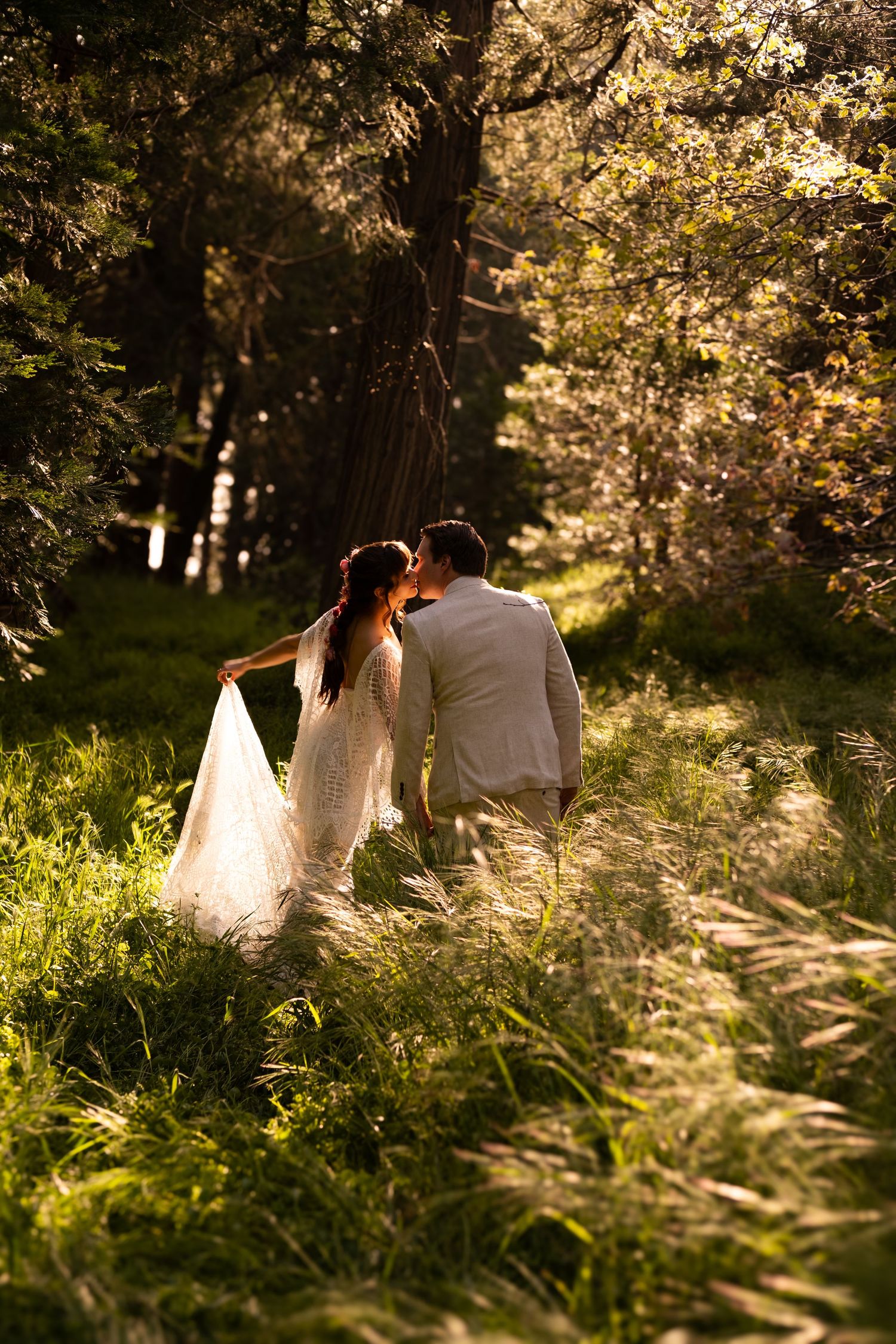 Bride and groom walking through a sun-drenched forest, bride in flowing white lace wedding gown.