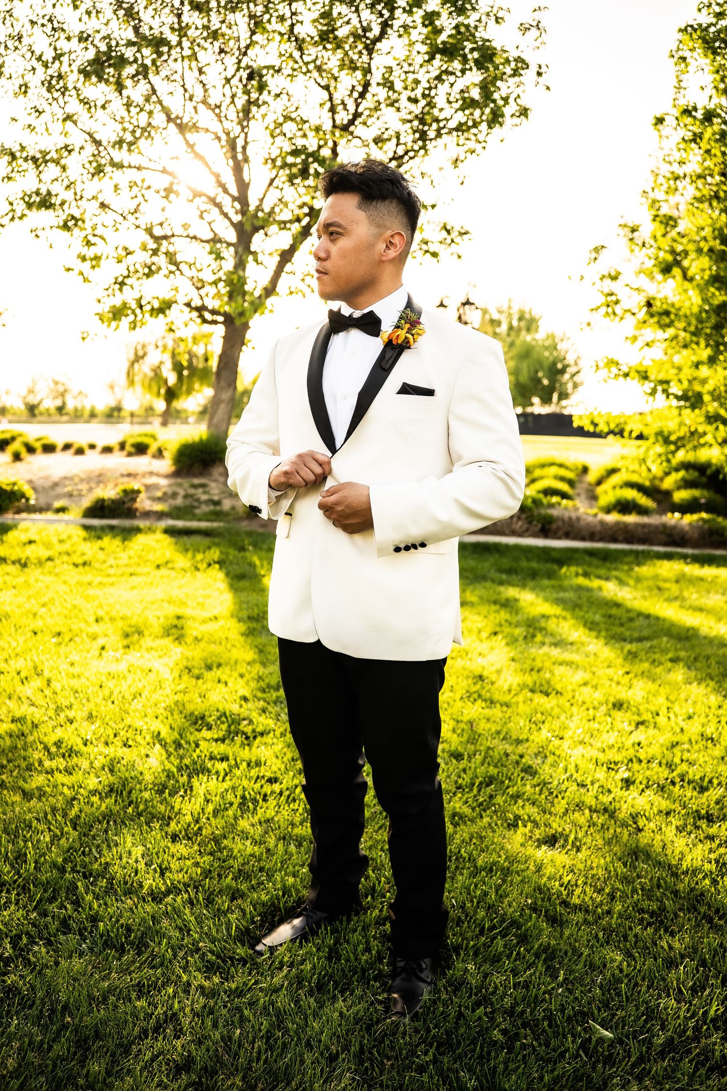 Elegant groom in white tuxedo jacket with black bow tie and boutonniere standing on green lawn at sunset.