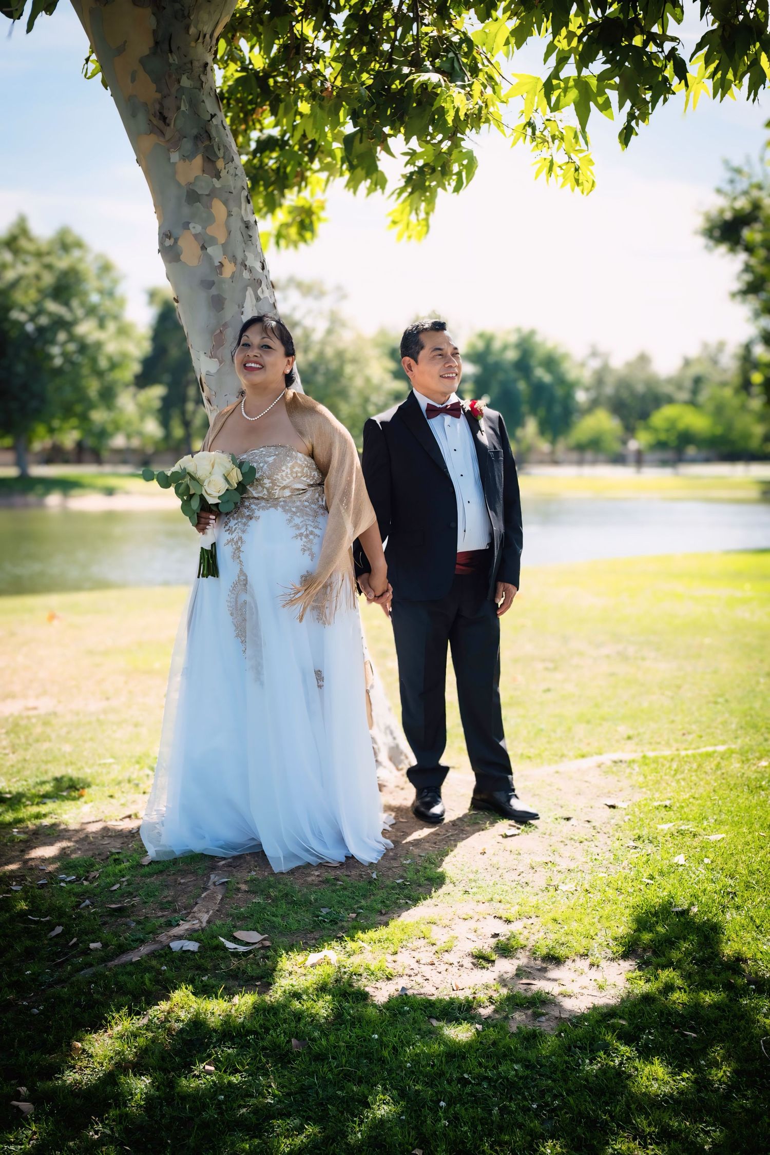 Bride and groom holding hands under a large tree beside a lake on their sunny wedding day.