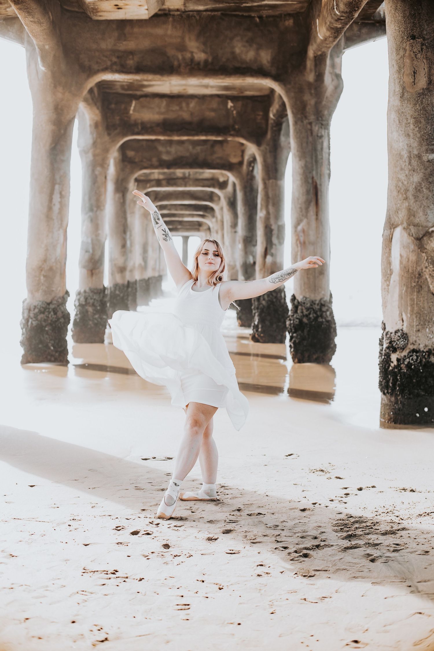 Young woman in white dress dancing gracefully under a beach pier on sandy shore.