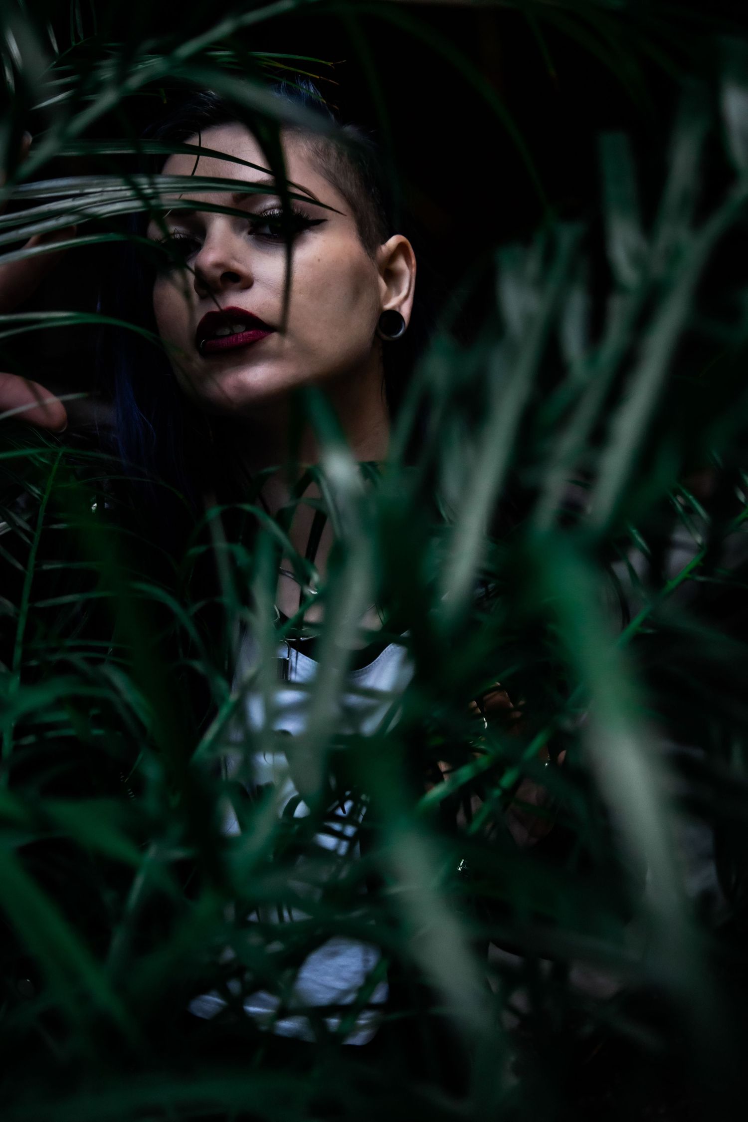 A woman with bold red lips and shaved head partially hidden among dark green tropical leaves.