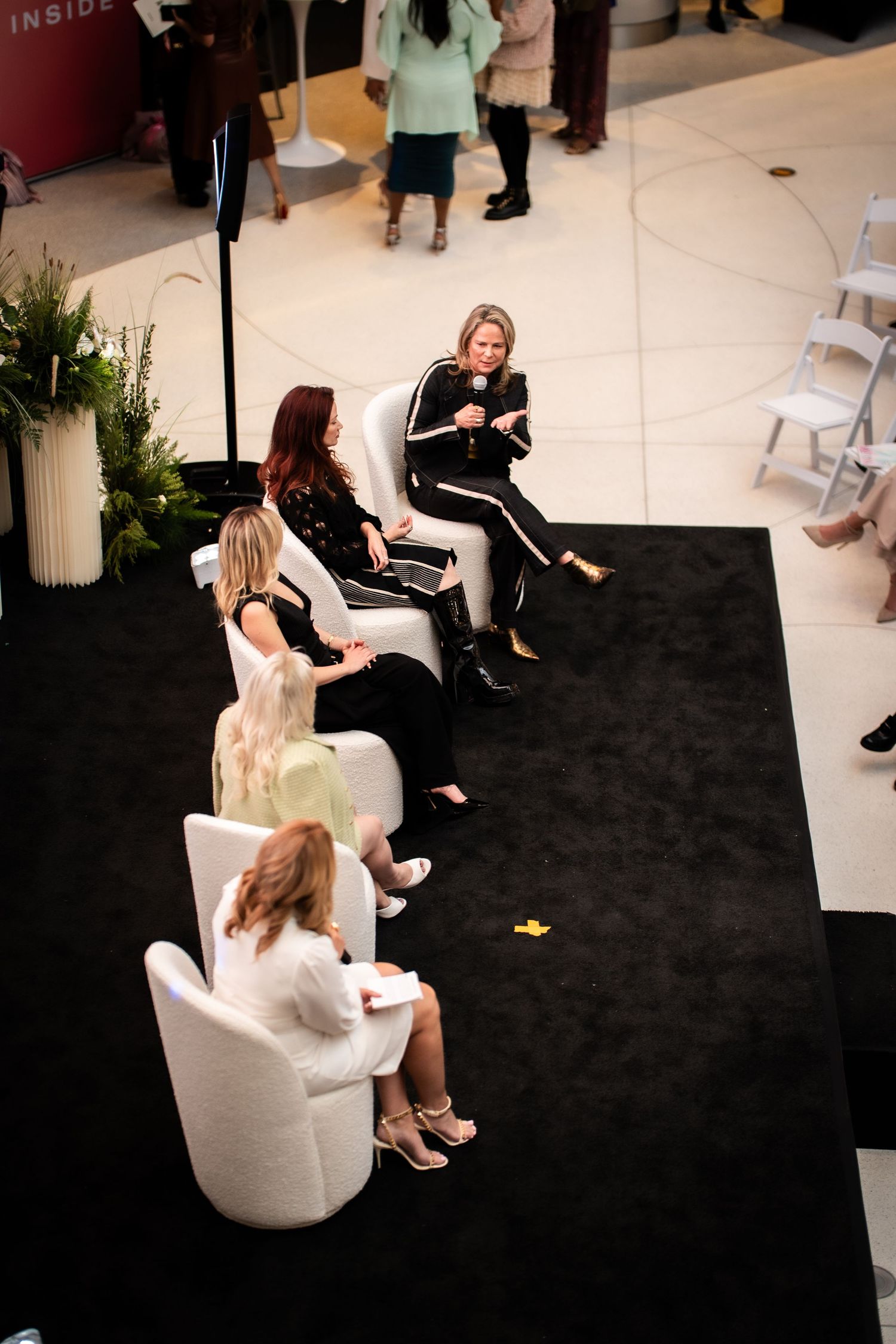 Panel discussion on a black stage with four women seated in white chairs at an indoor event.