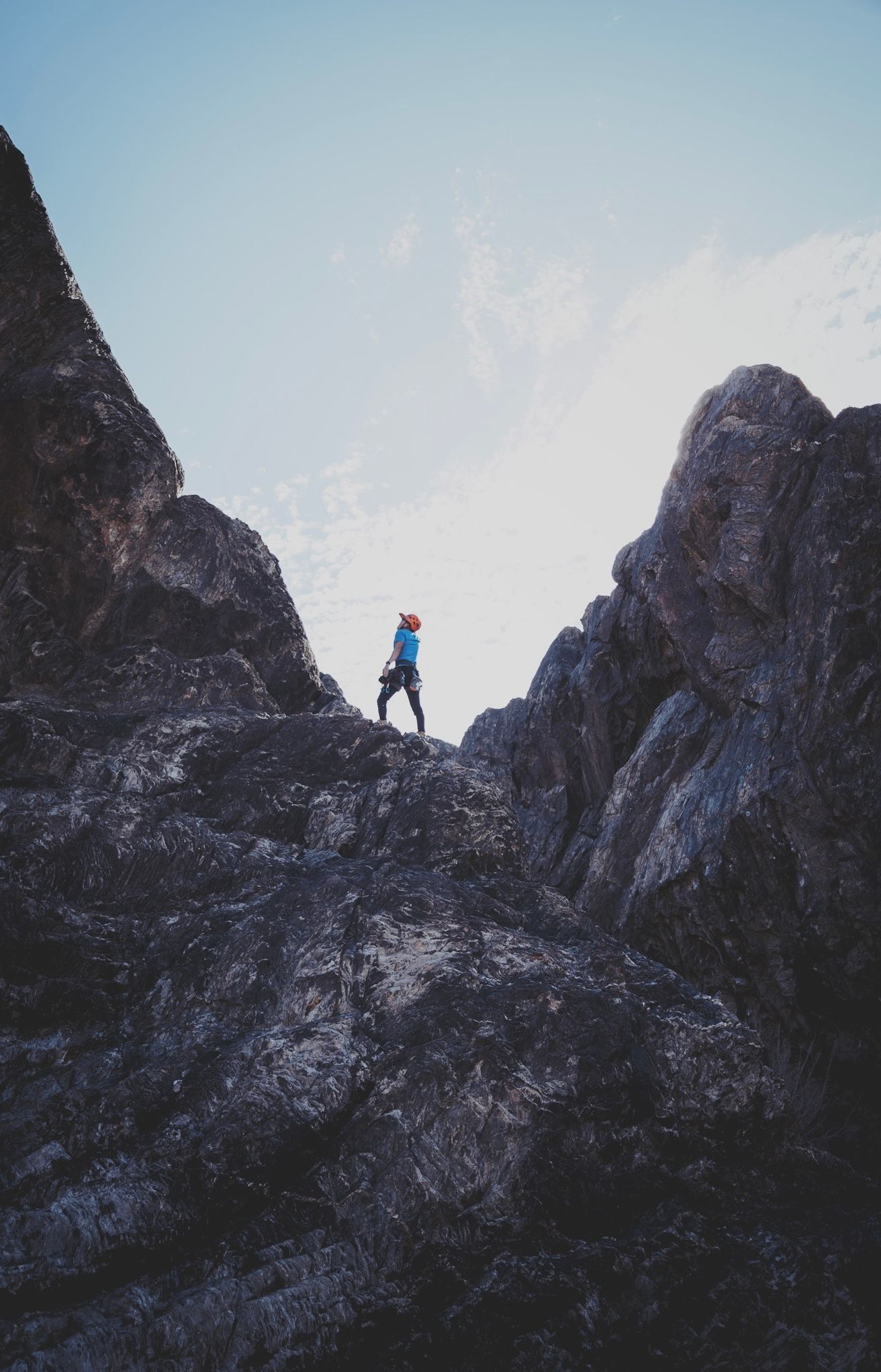 A lone climber stands on a rocky mountain ridge between two peaks against a clear blue sky.
