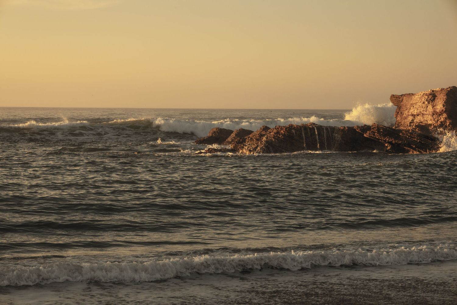 Waves crash against rocky coastal cliffs at golden sunset, with warm orange hues reflecting on the ocean surface.