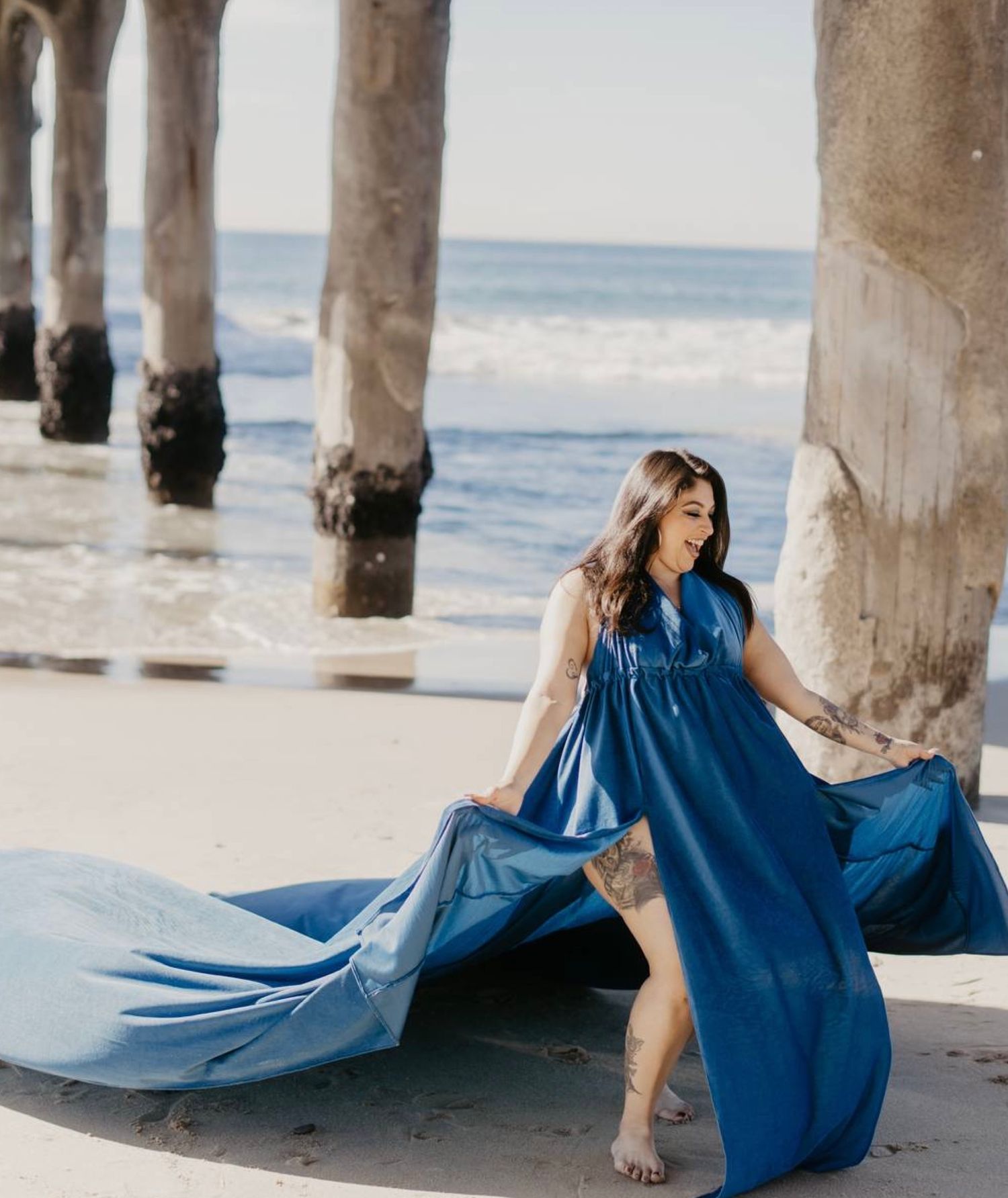 Woman in vibrant blue maxi dress laughing as fabric flows in wind near beach pier.