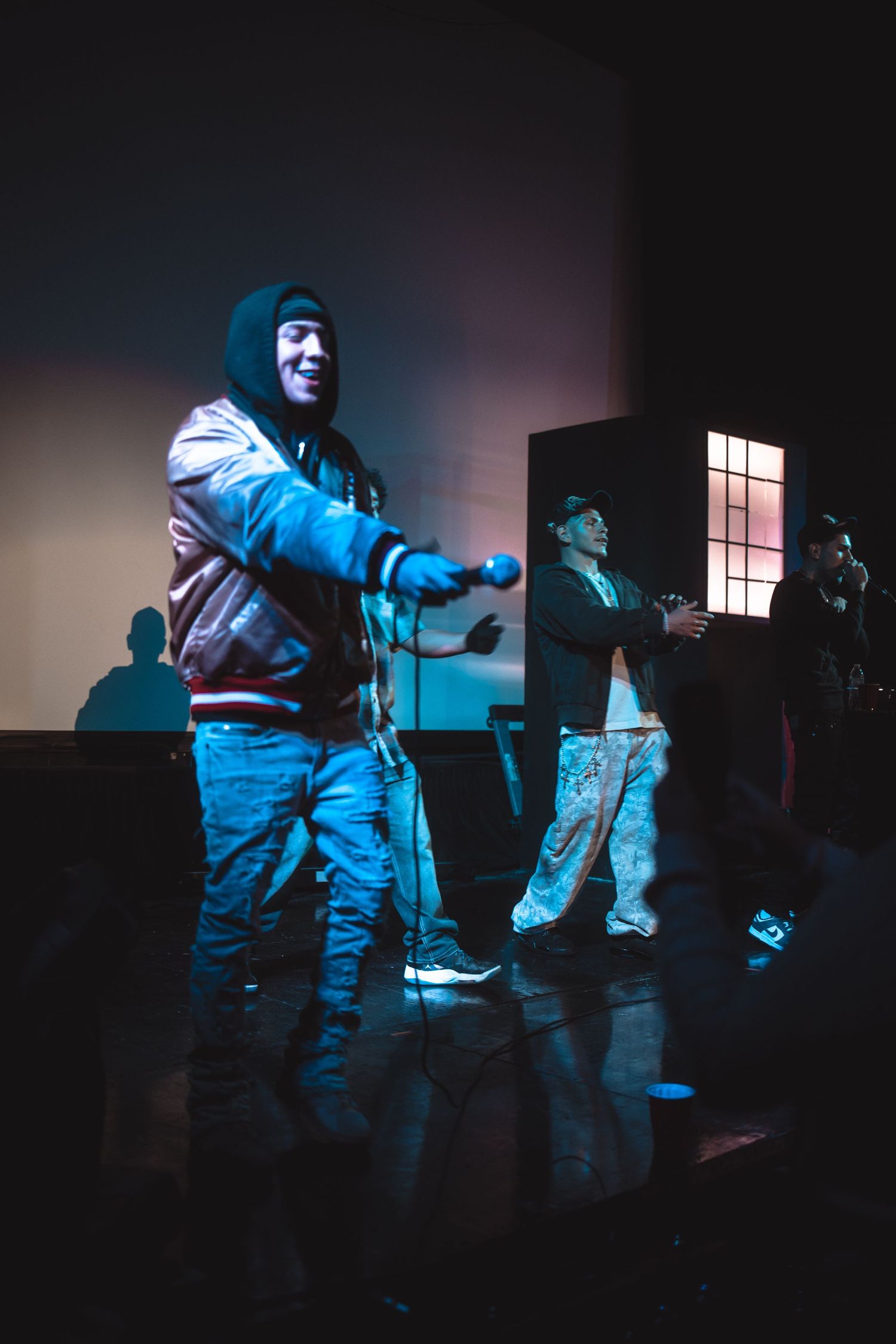 Hip-hop performer in a bomber jacket energetically rapping on stage under blue lighting with bandmates behind him.