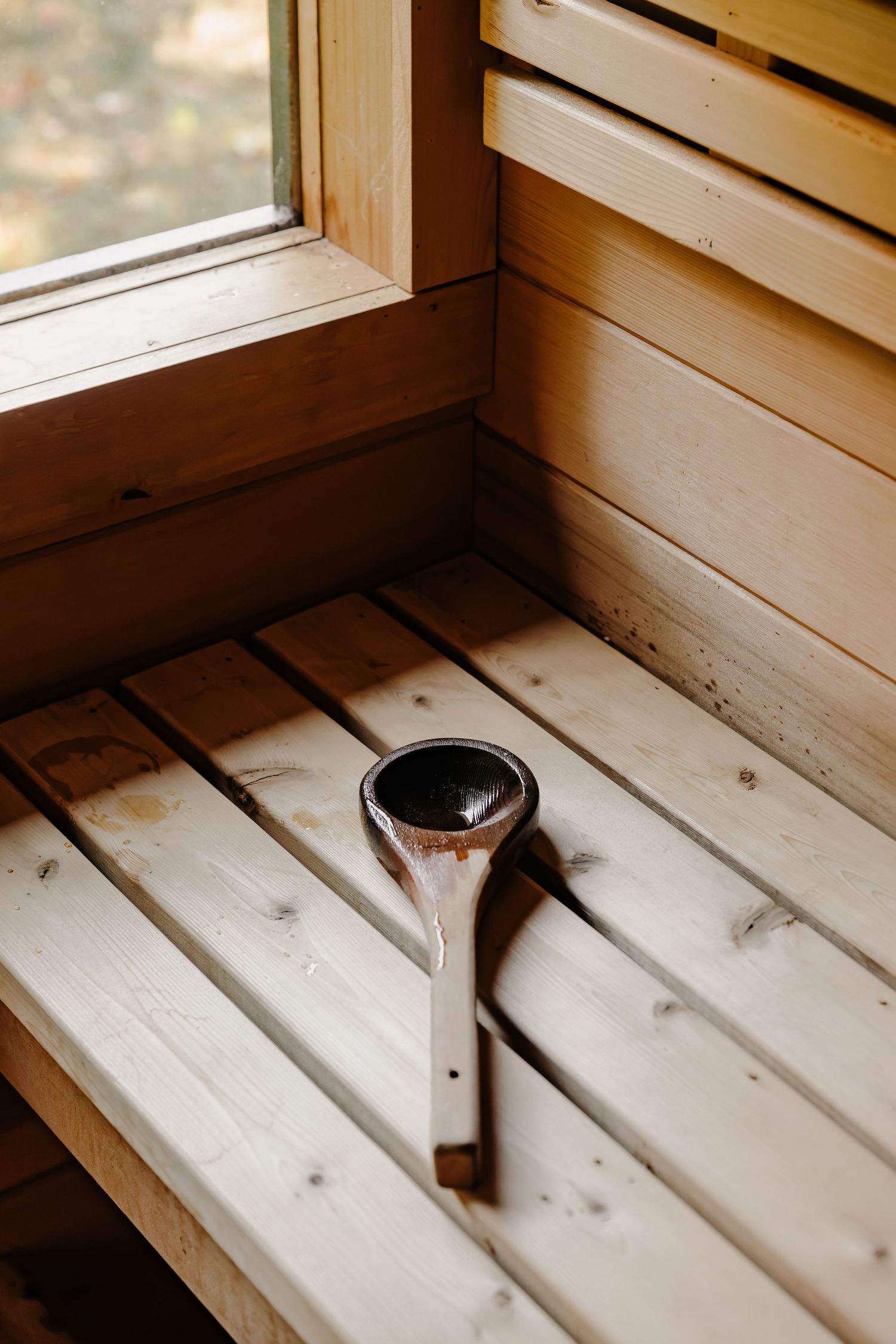 Wooden sauna ladle resting on light pine slatted benches beside a window in a cozy traditional Finnish sauna.