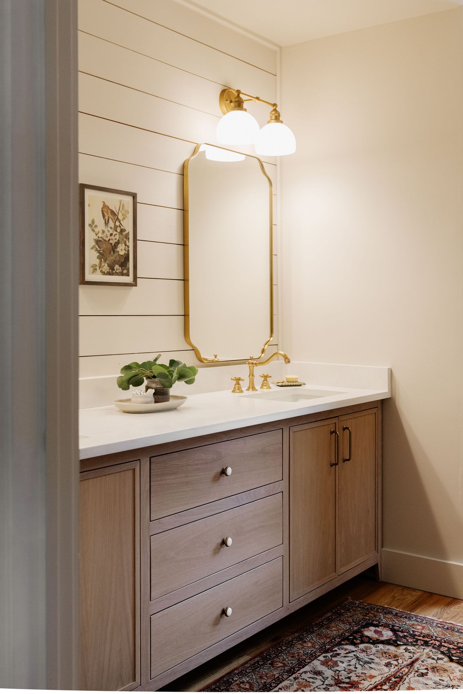Elegant bathroom vanity with wood cabinets, gold fixtures, shiplap wall, brass mirror, and vintage rug on hardwood floors.
