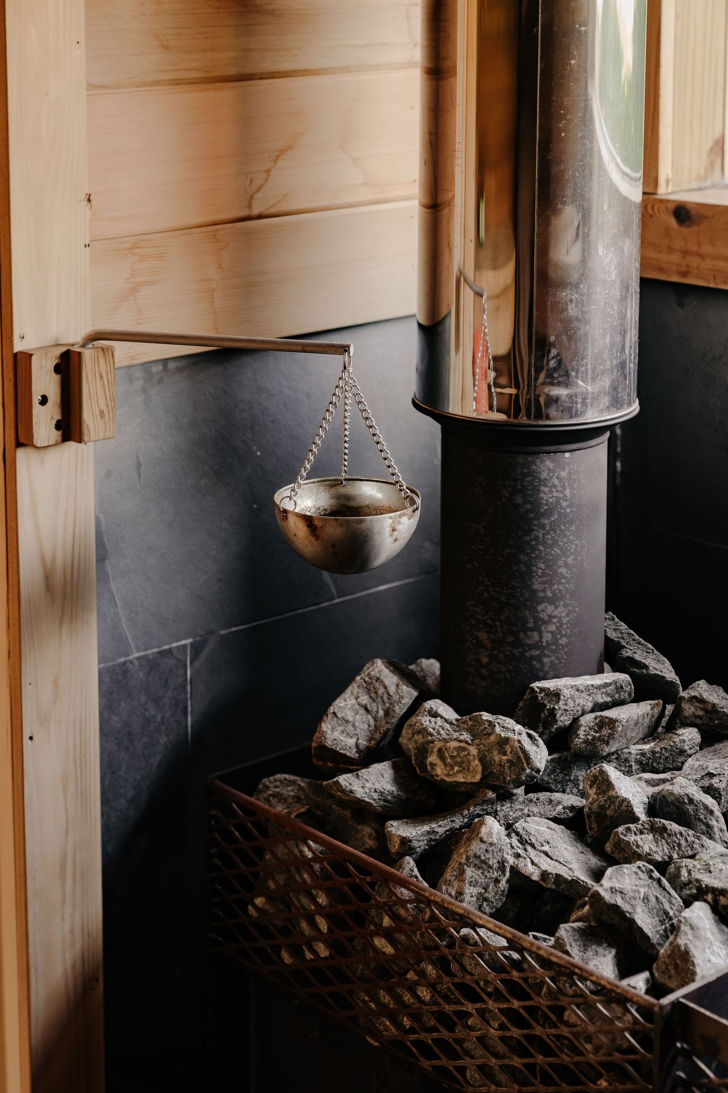 Sauna stove with a pile of heated stones, a hanging metal water ladle and a stainless steel chimney against dark slate and wood panels.