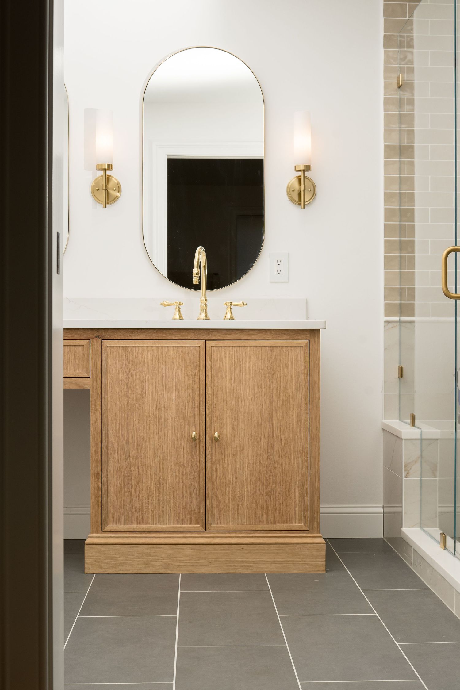 Modern bathroom featuring gray subway tile, brass sconces, and wooden vanity with round mirrors.