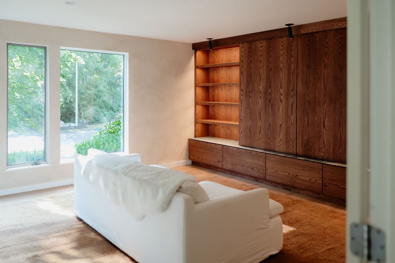 Modern living room with white sofa, wood herringbone floor, large windows, and built-in walnut shelving unit.