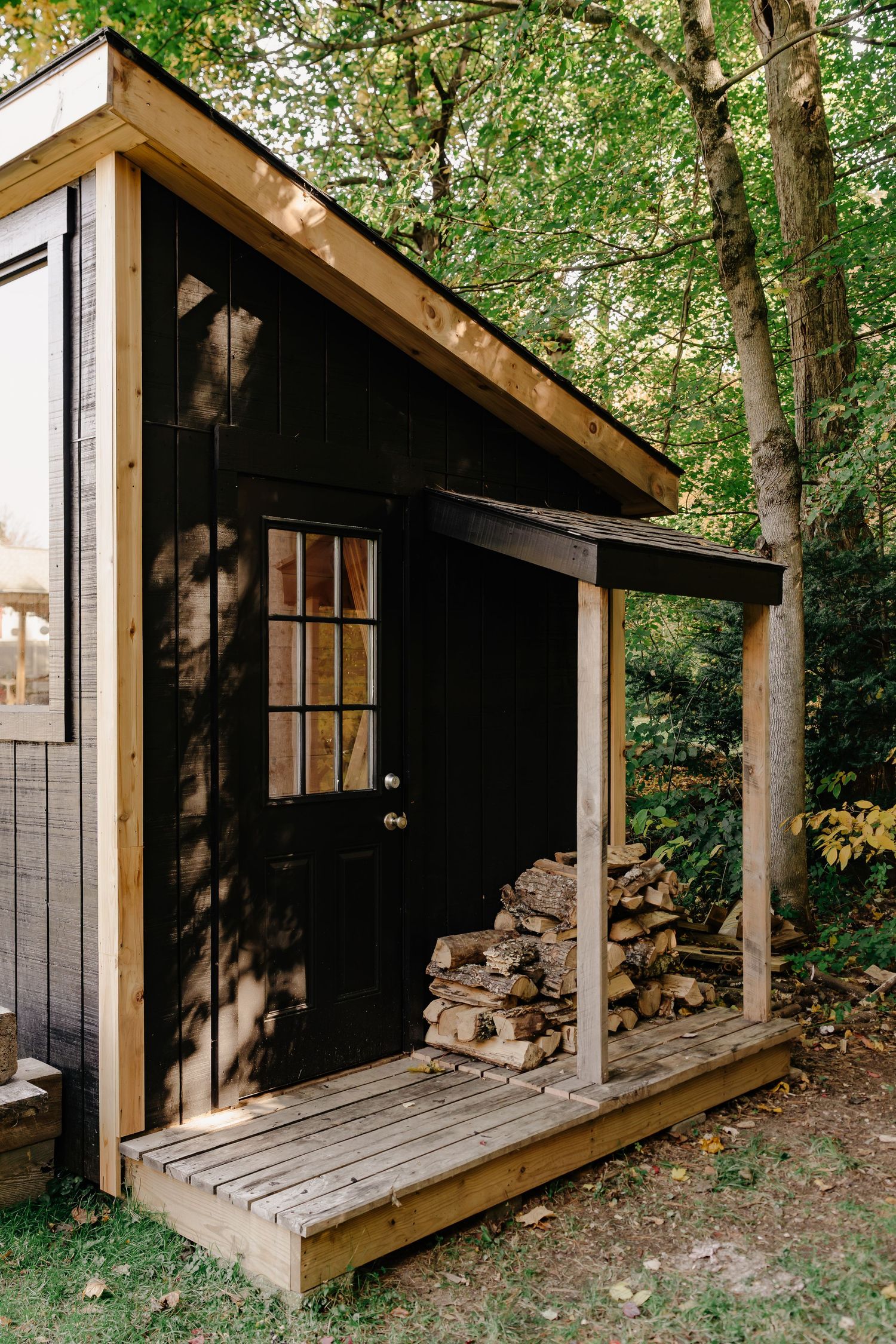 Rustic black cabin with a wooden porch, glass-paned door, and neatly stacked firewood beneath a covered overhang surrounded by trees.