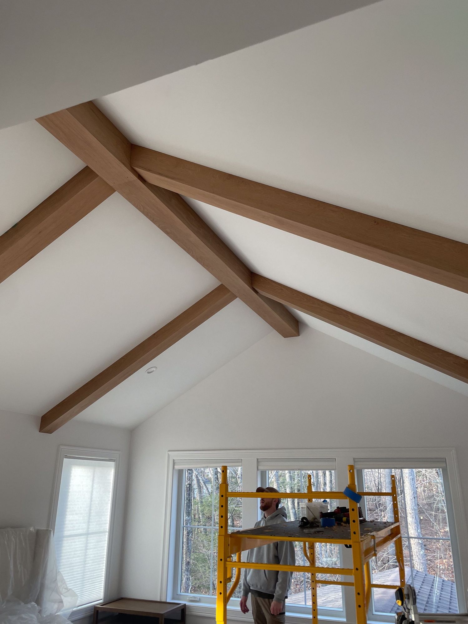 Room renovation showing exposed wooden ceiling beams and large windows with construction equipment.