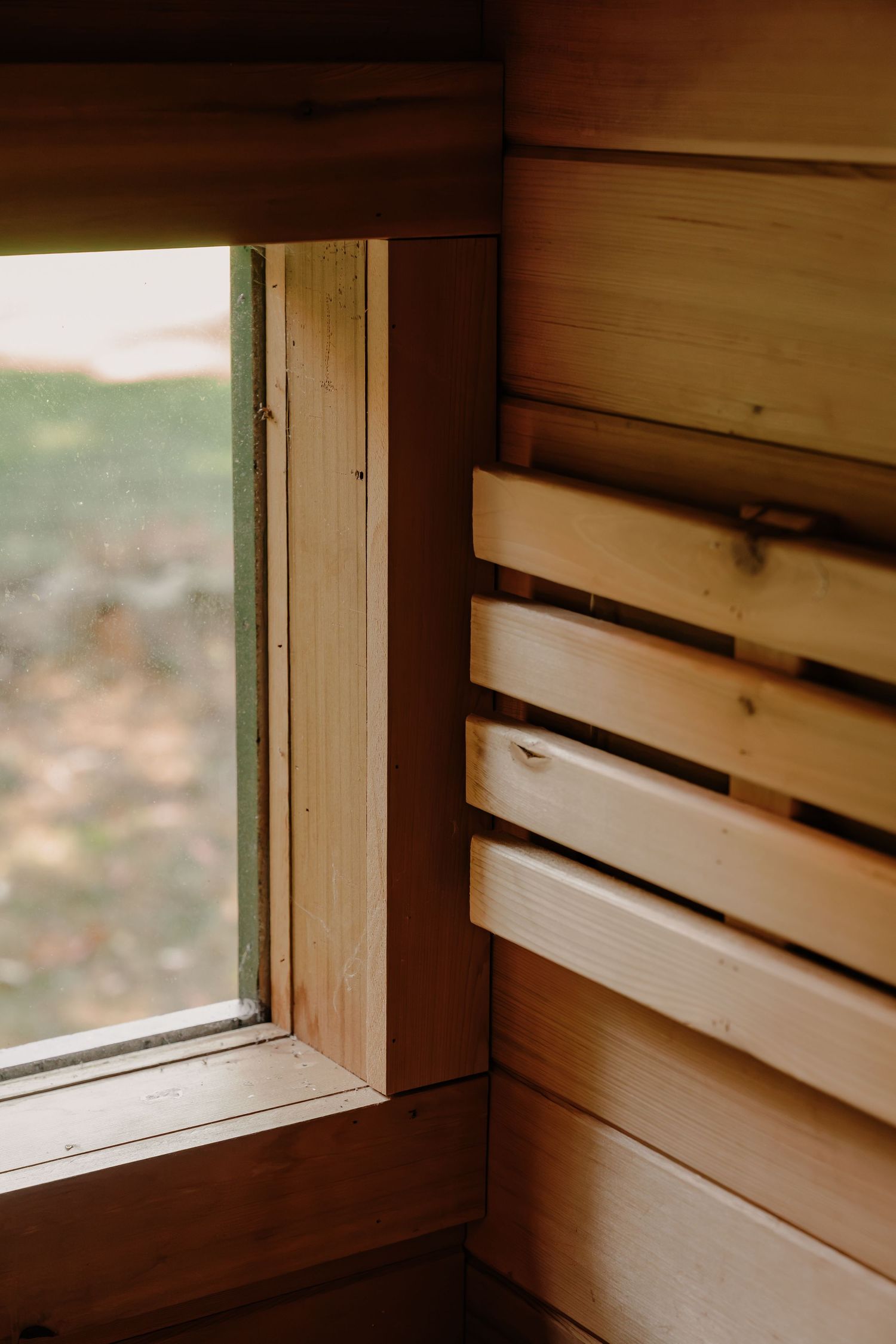 Sunlit window framed in natural cedar wood beside slatted wooden bench boards inside a cozy sauna cabin.