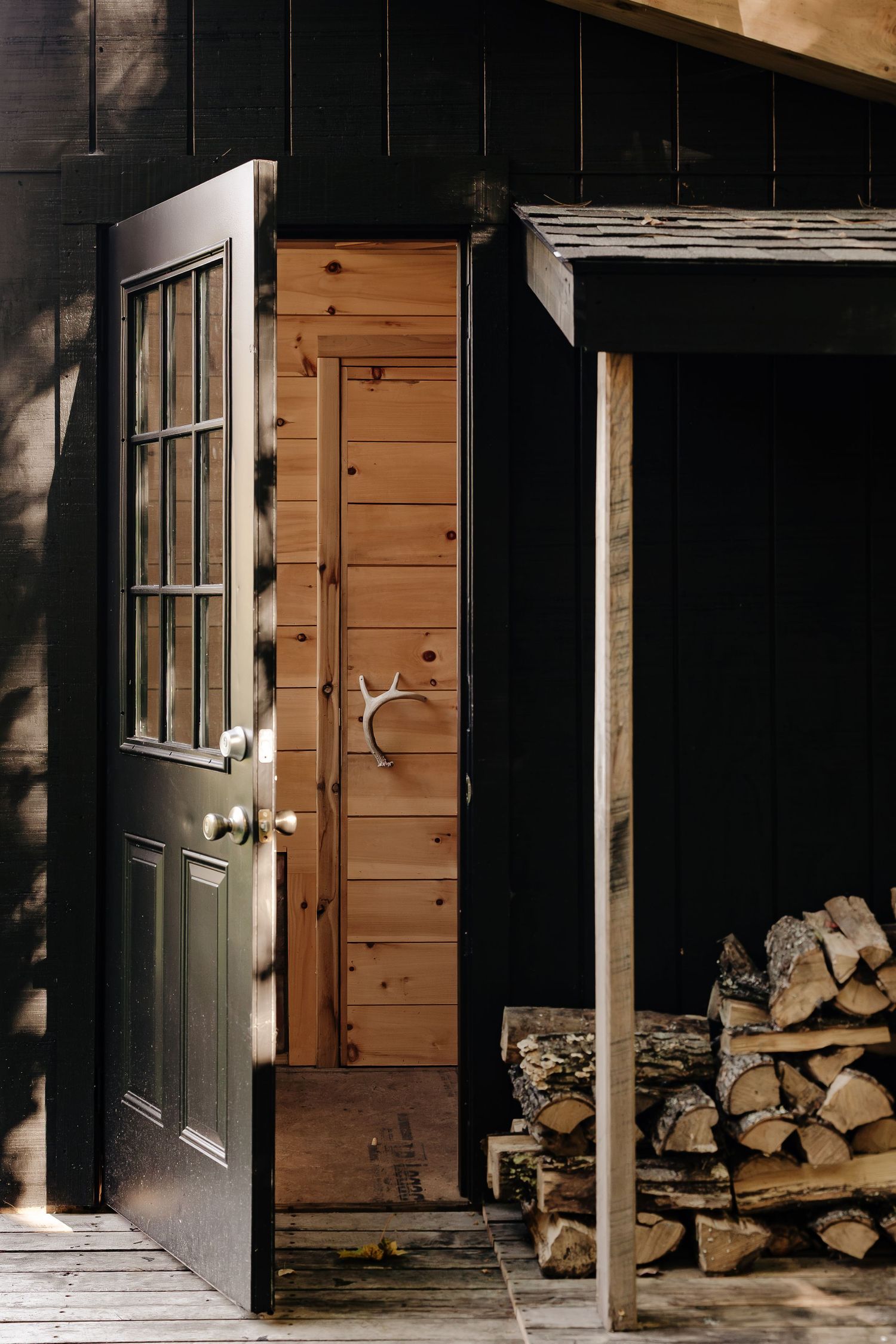 Open black door beside a knotty pine door with antler handle, next to a covered stack of split firewood on a wooden porch.