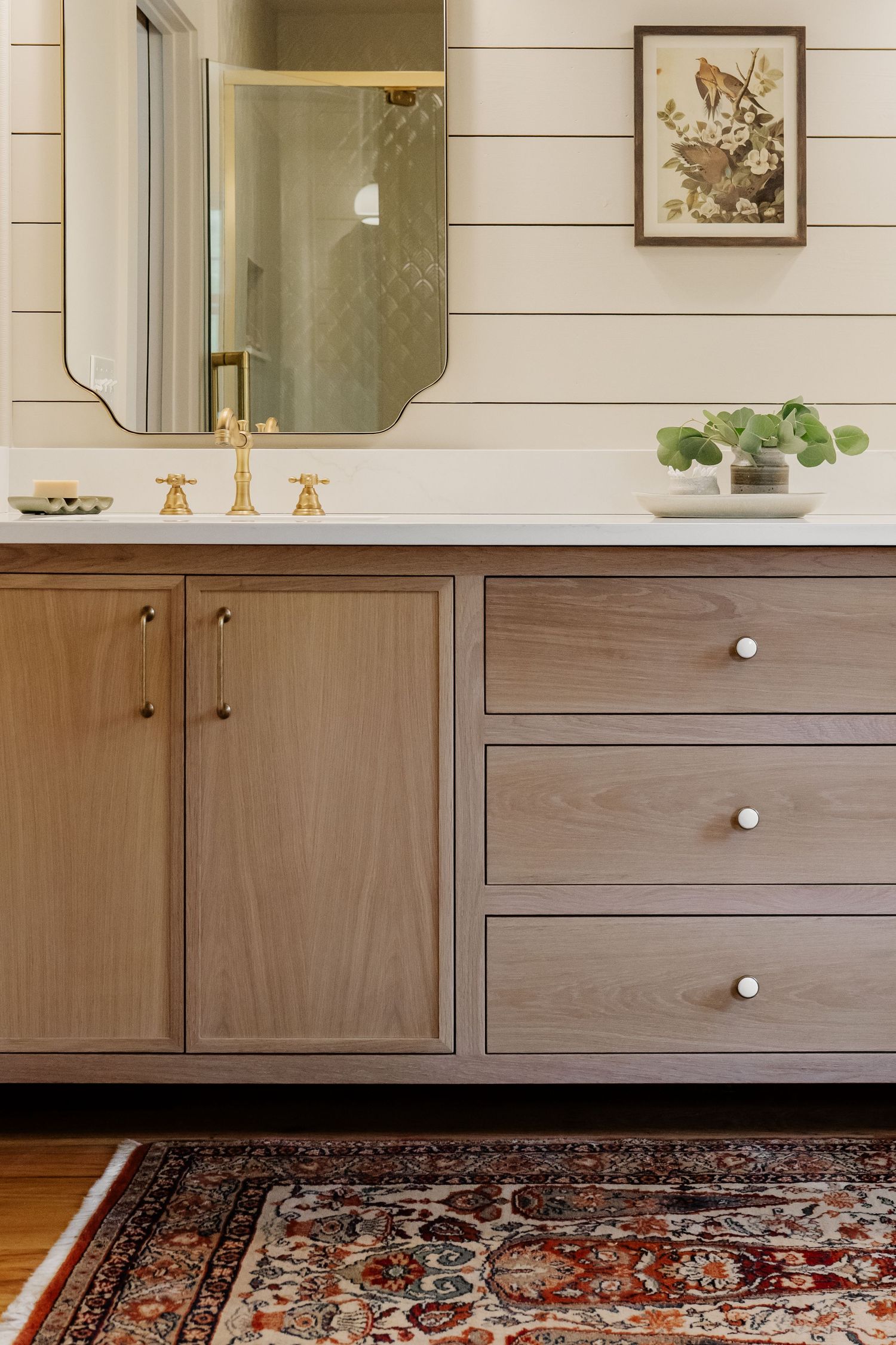 Elegant bathroom vanity with light wood cabinets, brass fixtures, ornate mirror, and a vintage Persian rug on hardwood floors.