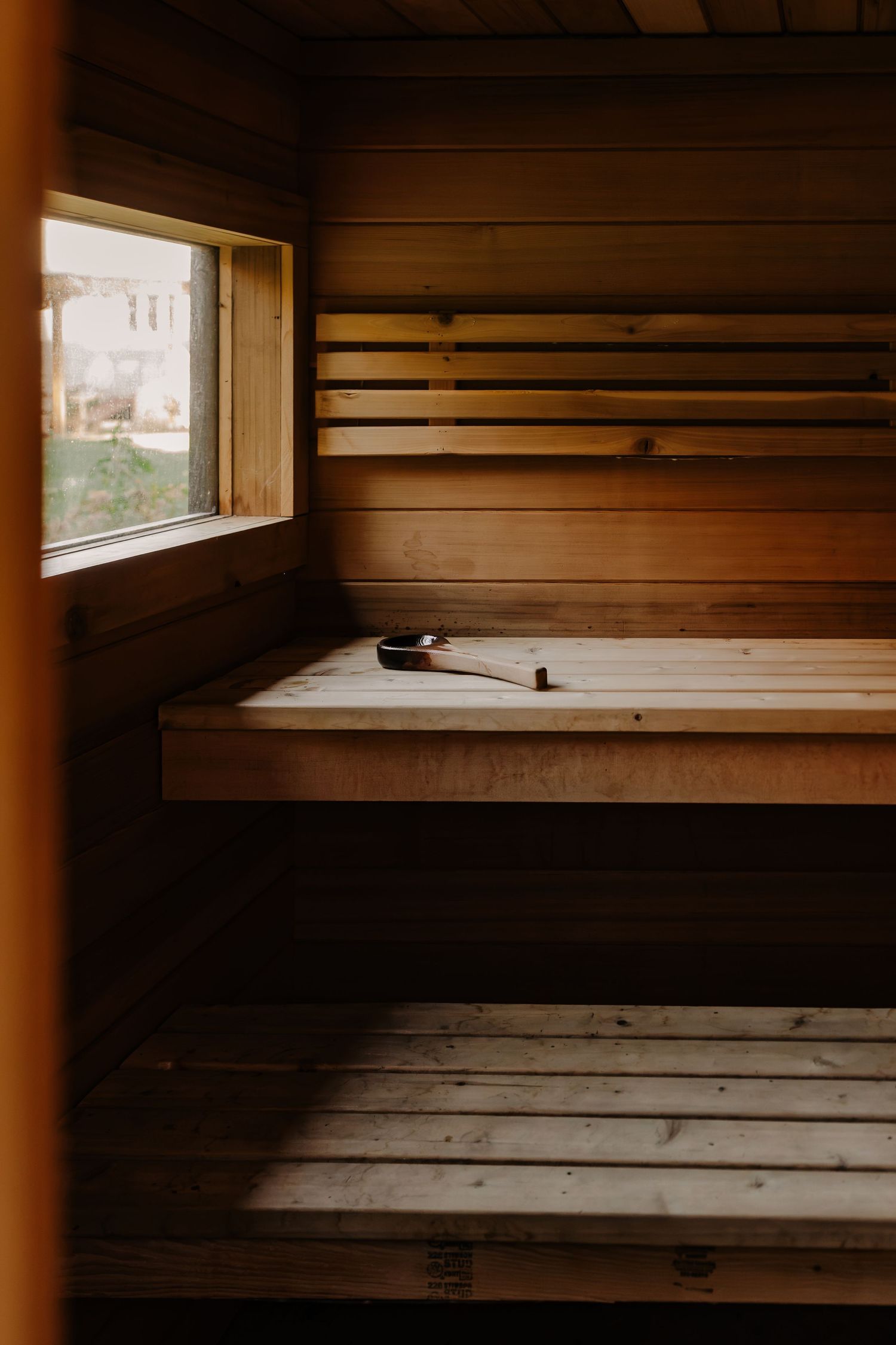 Cedar wood sauna interior with tiered benches, a wooden ladle resting on the upper bench, and a small window letting in soft natural light.