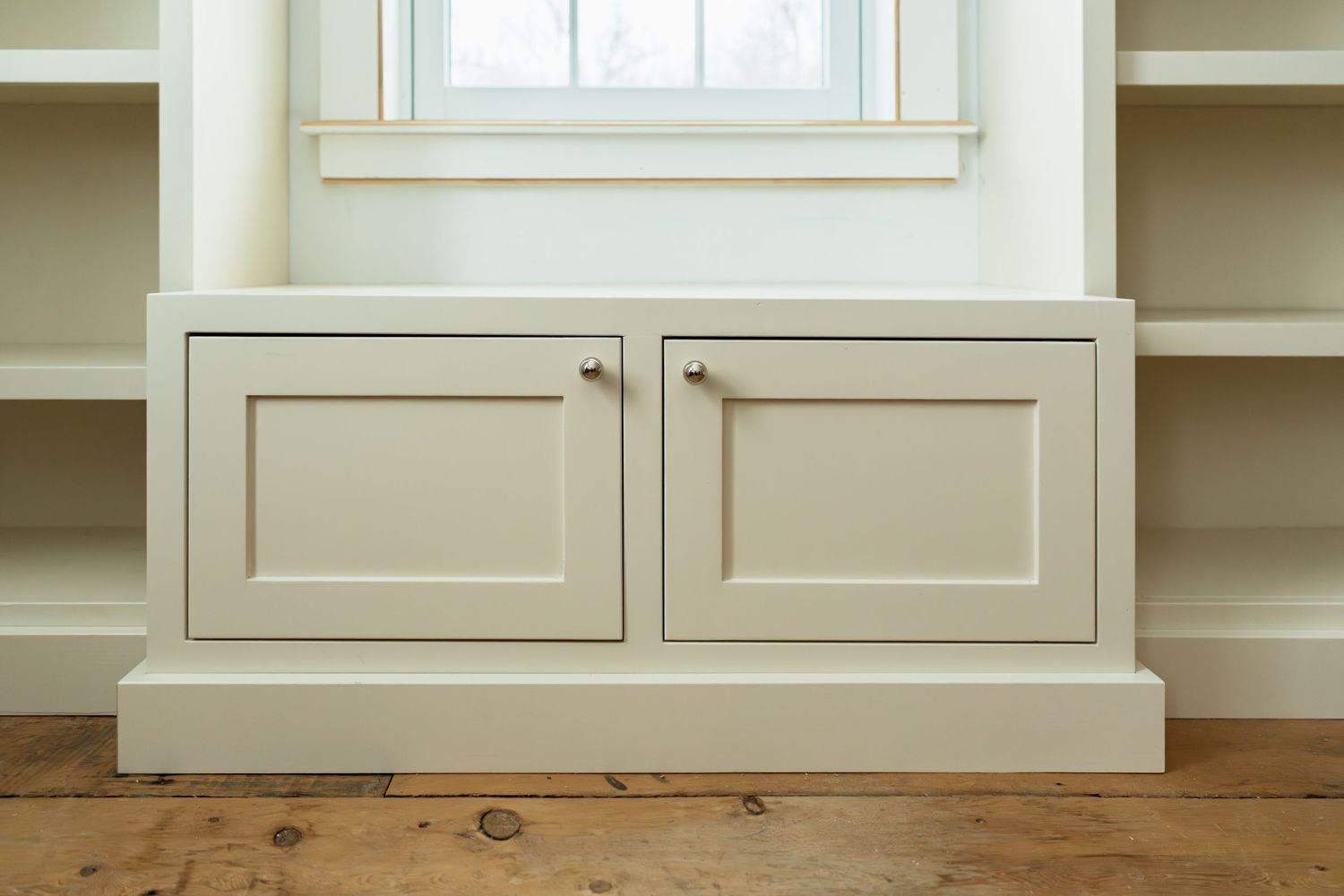 Custom white built-in shelving unit with cabinet doors and rustic wood flooring in a bright room.