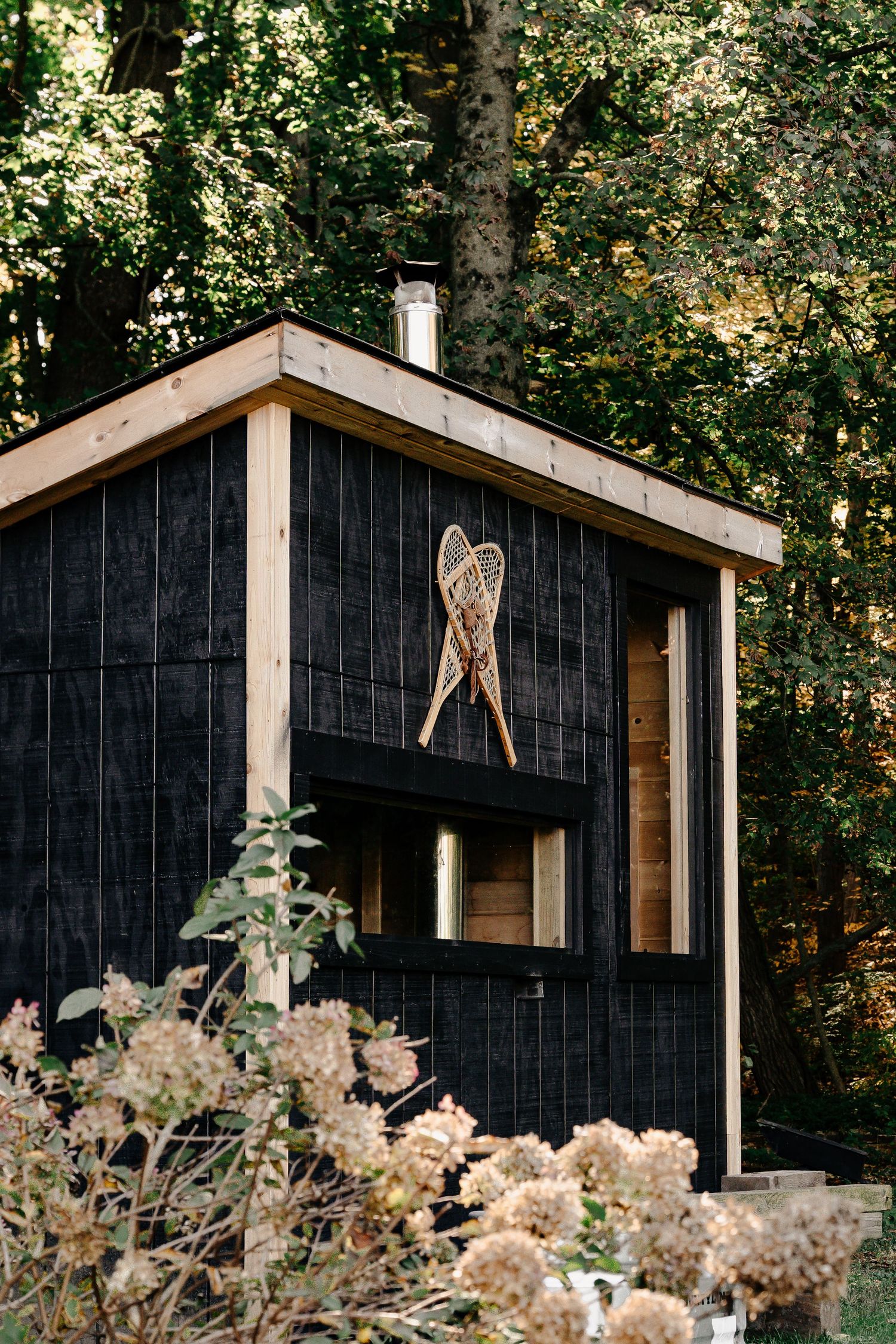 Black wood-clad sauna cabin with a chimney and vintage snowshoes mounted on the wall, surrounded by trees and blooming hydrangeas.