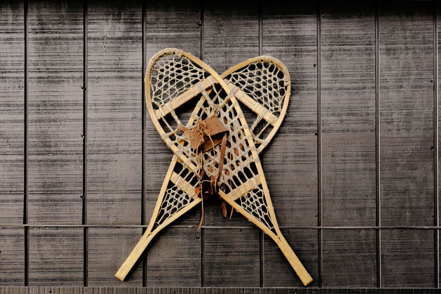 Pair of vintage wooden snowshoes with rawhide lacing and leather bindings crossed and hung on a weathered gray plank wall.