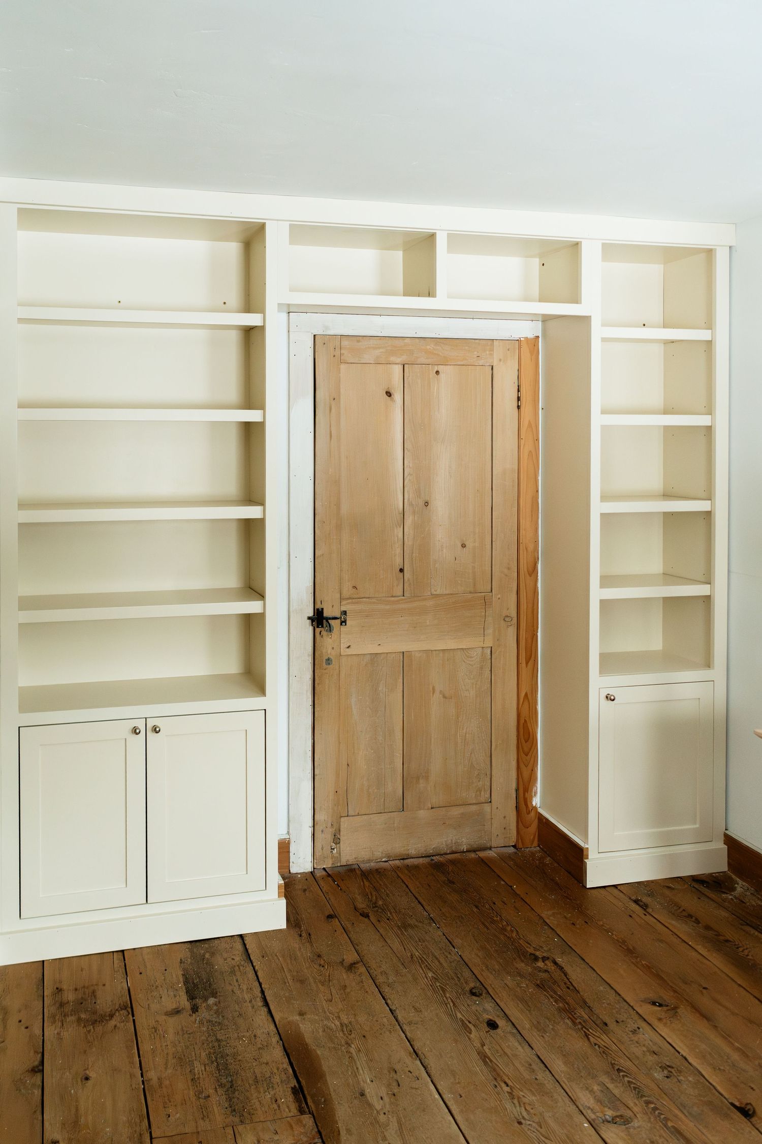 Floor-to-ceiling built-in storage featuring white shelving units and a natural wood door on rustic flooring.
