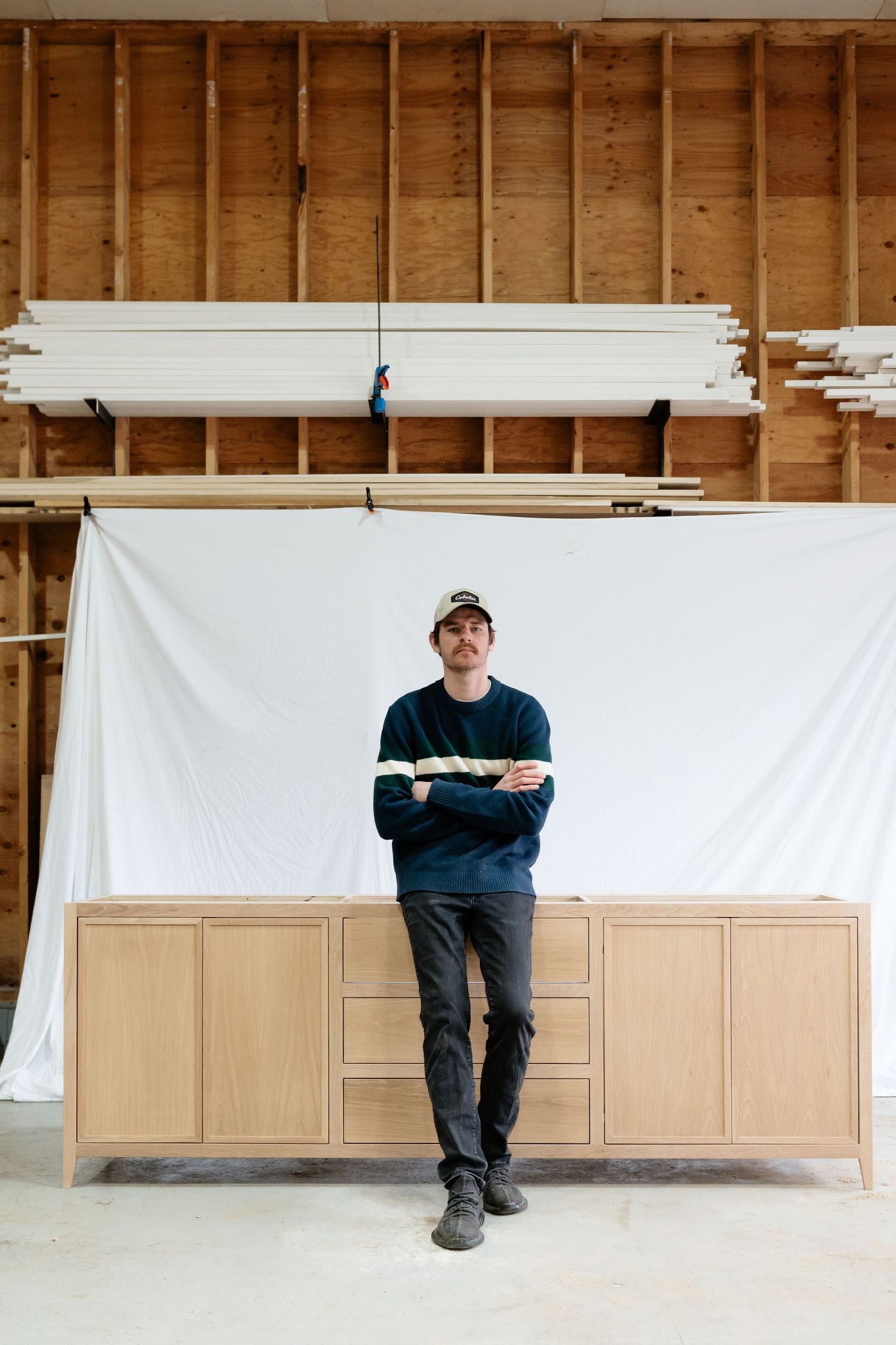 Man in navy sweater and cap leans against a light wood credenza in a workshop with lumber stacked behind him.