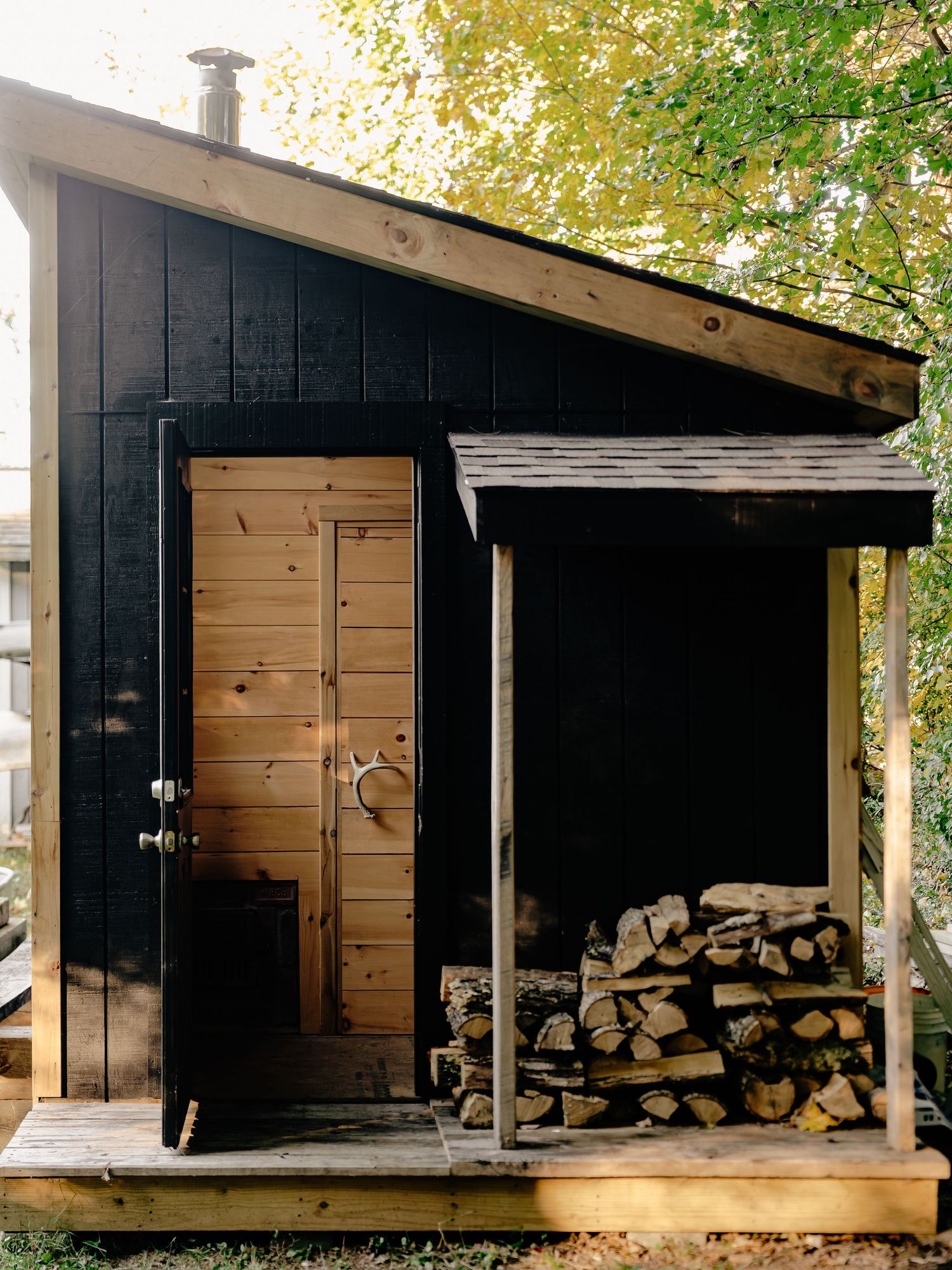 Modern black cedar sauna cabin with a slanted roof, natural wood door, and neatly stacked firewood under a covered porch.