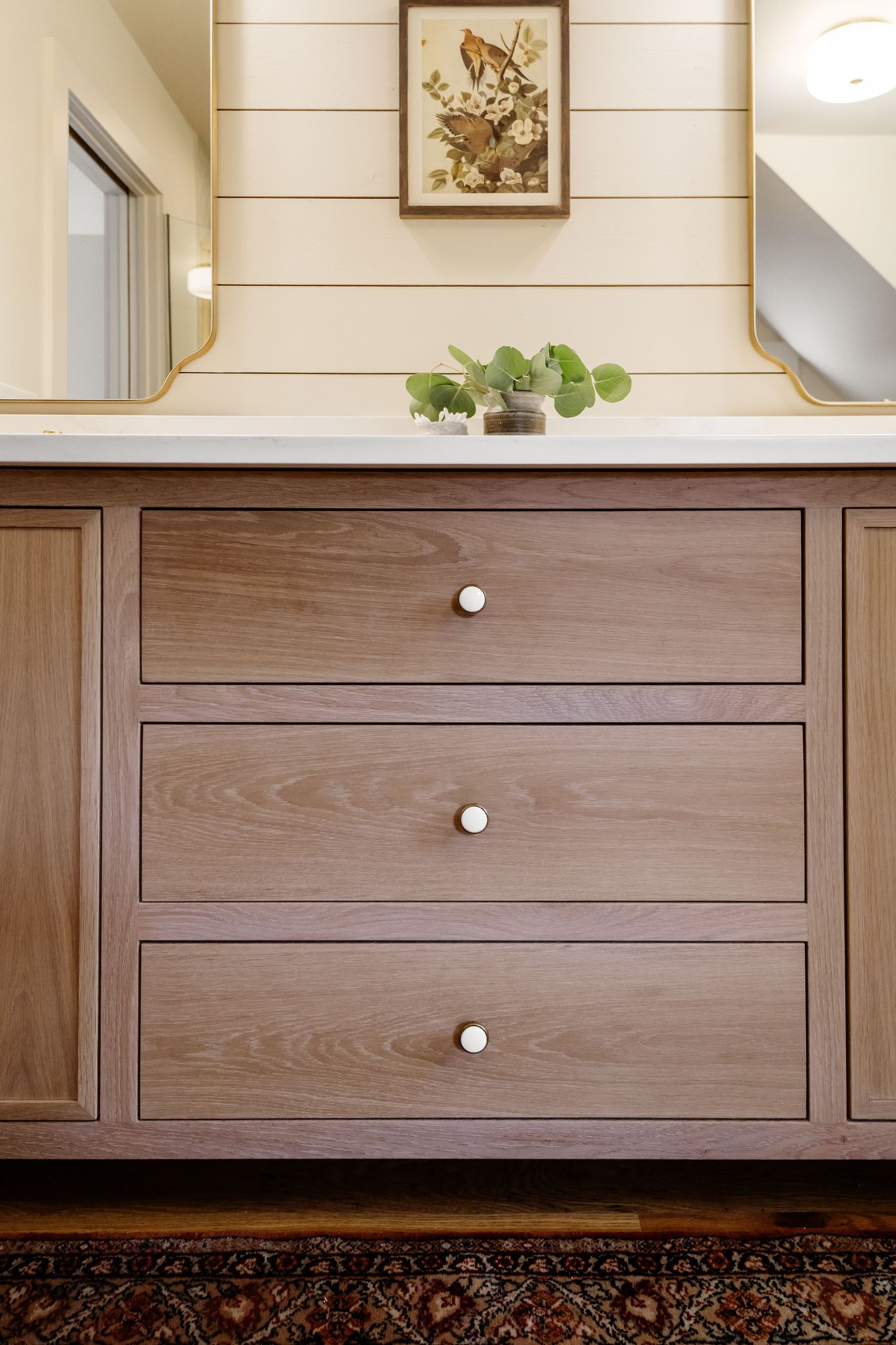 Wooden bathroom vanity with three drawers, marble countertop, dual mirrors, and botanical art on shiplap wall.