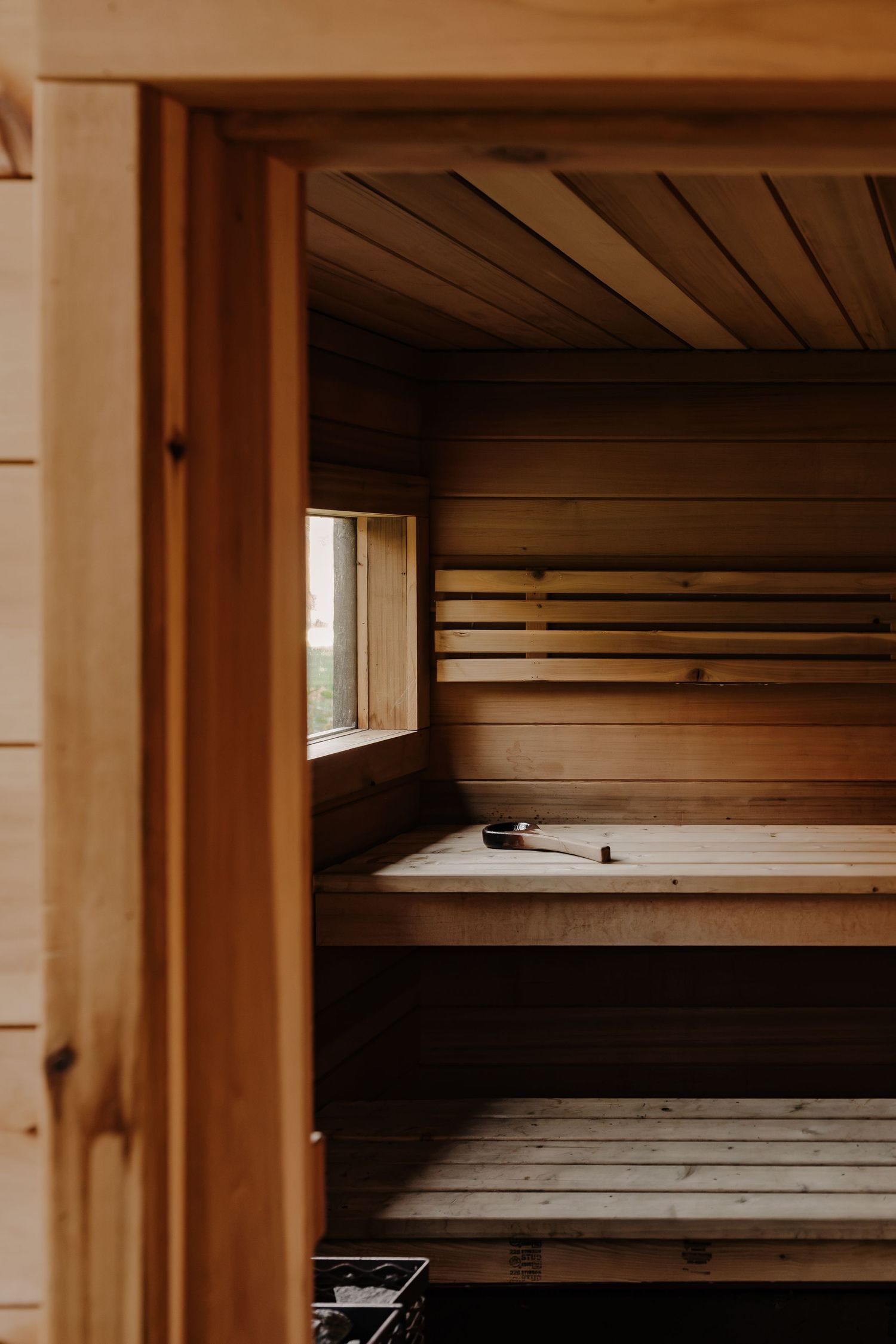 Empty cedar wood sauna interior with tiered benches, a small window, a wooden ladle, and warm natural light through an open door.