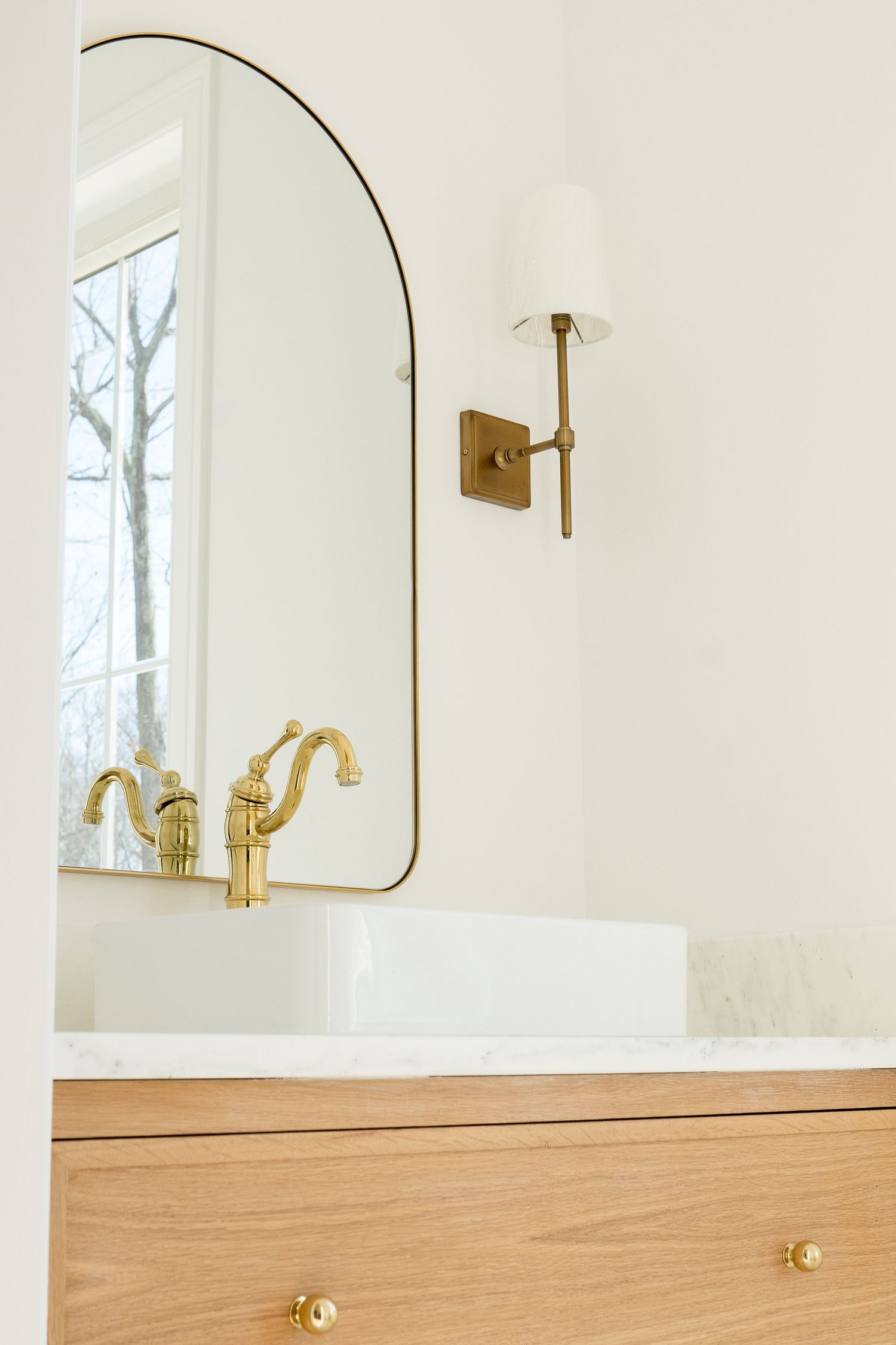 Close-up of oval mirror with brass trim and wall sconce above wooden bathroom vanity.