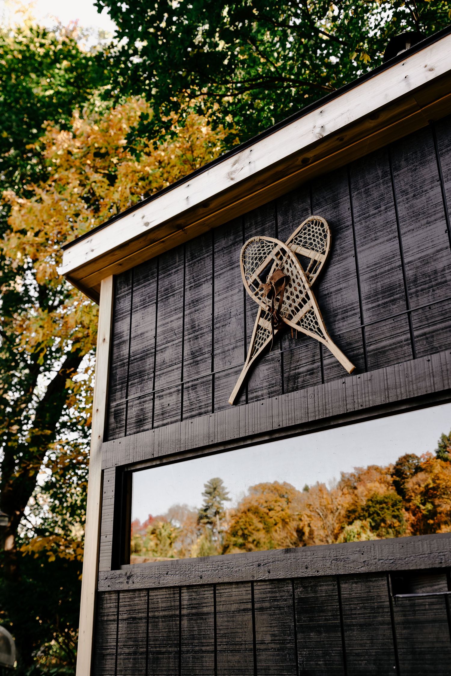 Pair of vintage wooden snowshoes mounted on a dark cabin wall, with a window reflecting colorful autumn foliage.