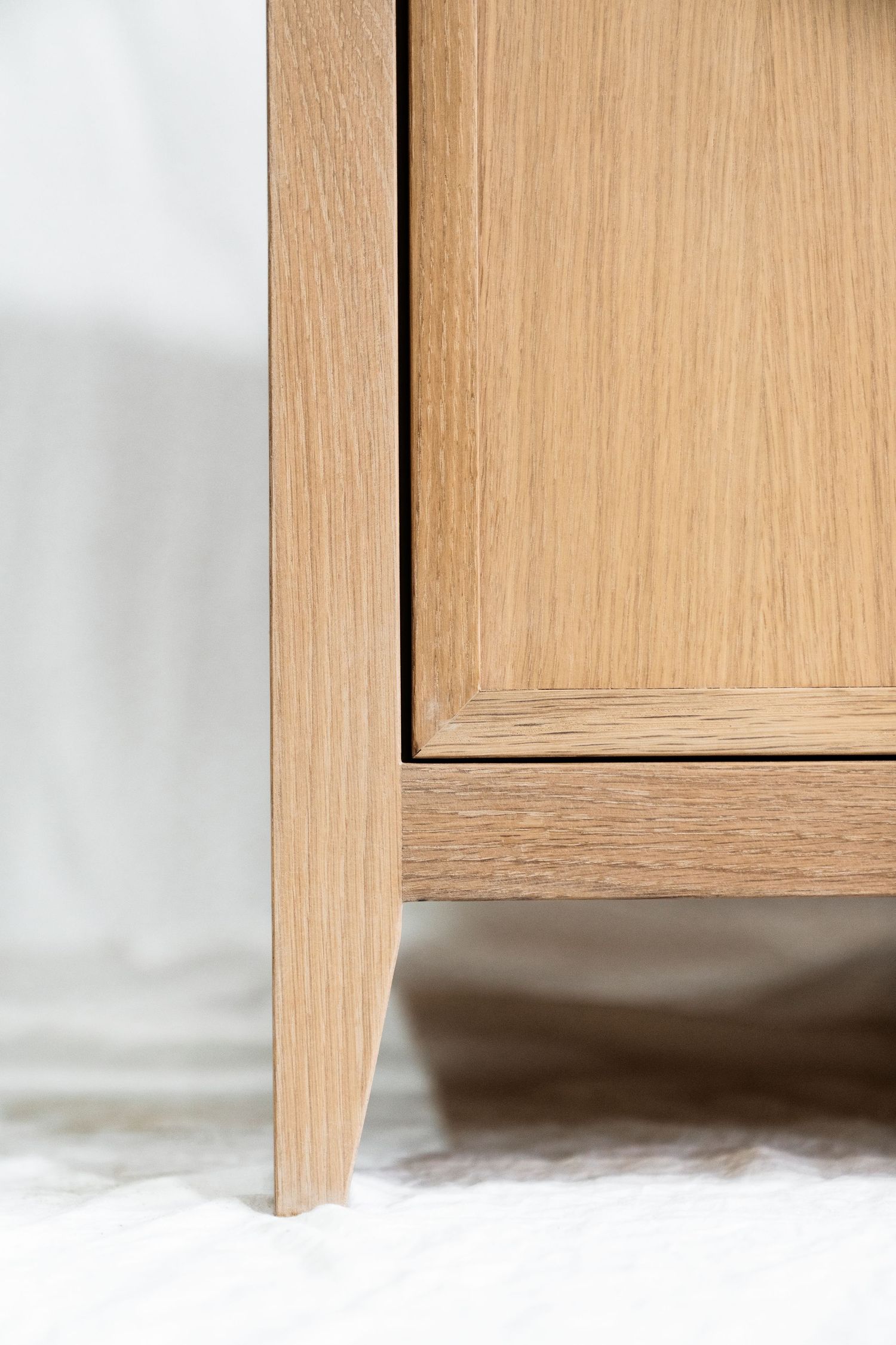 Close-up of a light oak wood cabinet corner with tapered leg on a white fluffy rug, showcasing natural wood grain.