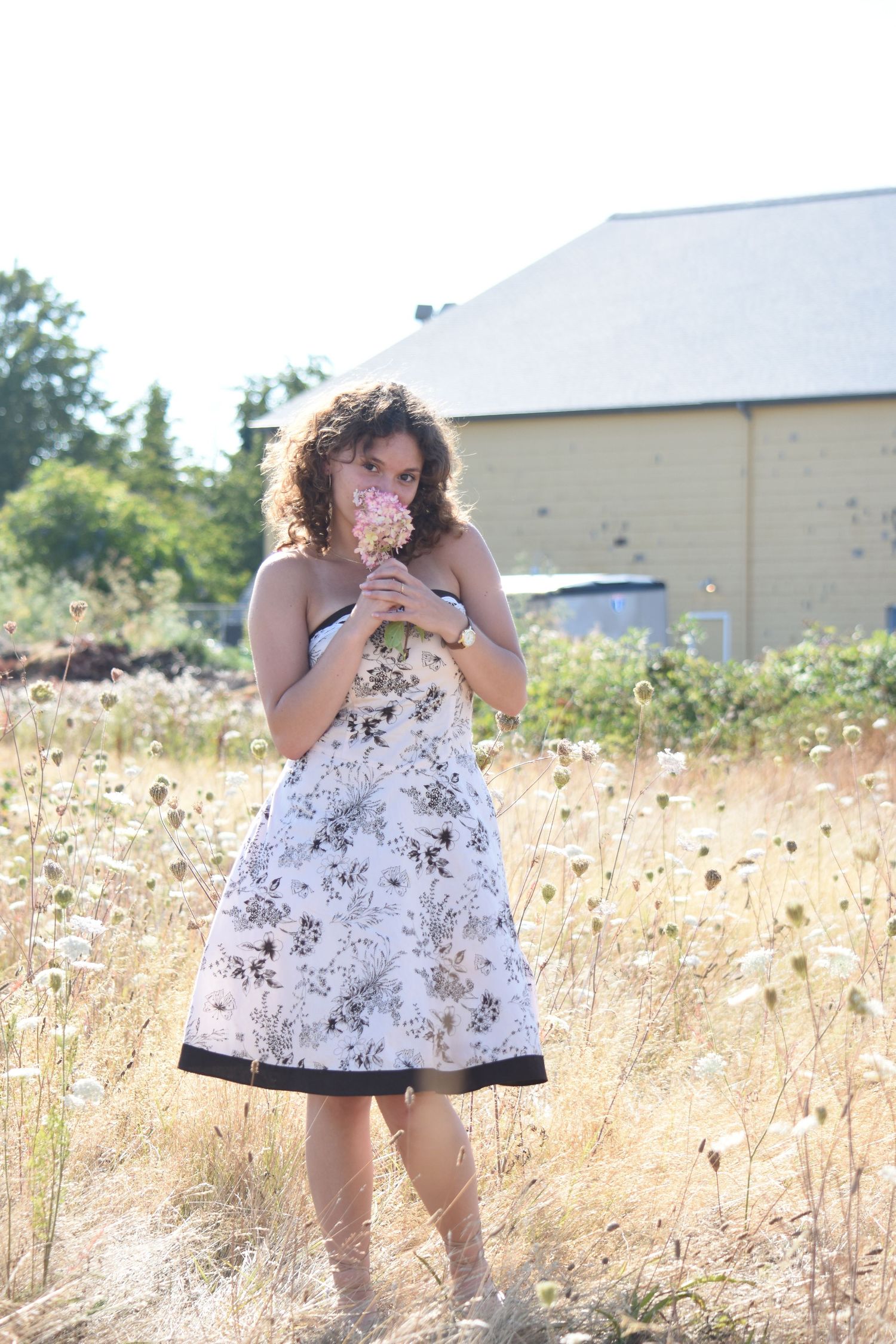 A person wearing a floral sundress stands in a golden field of tall grass and wildflowers in summer sunlight.