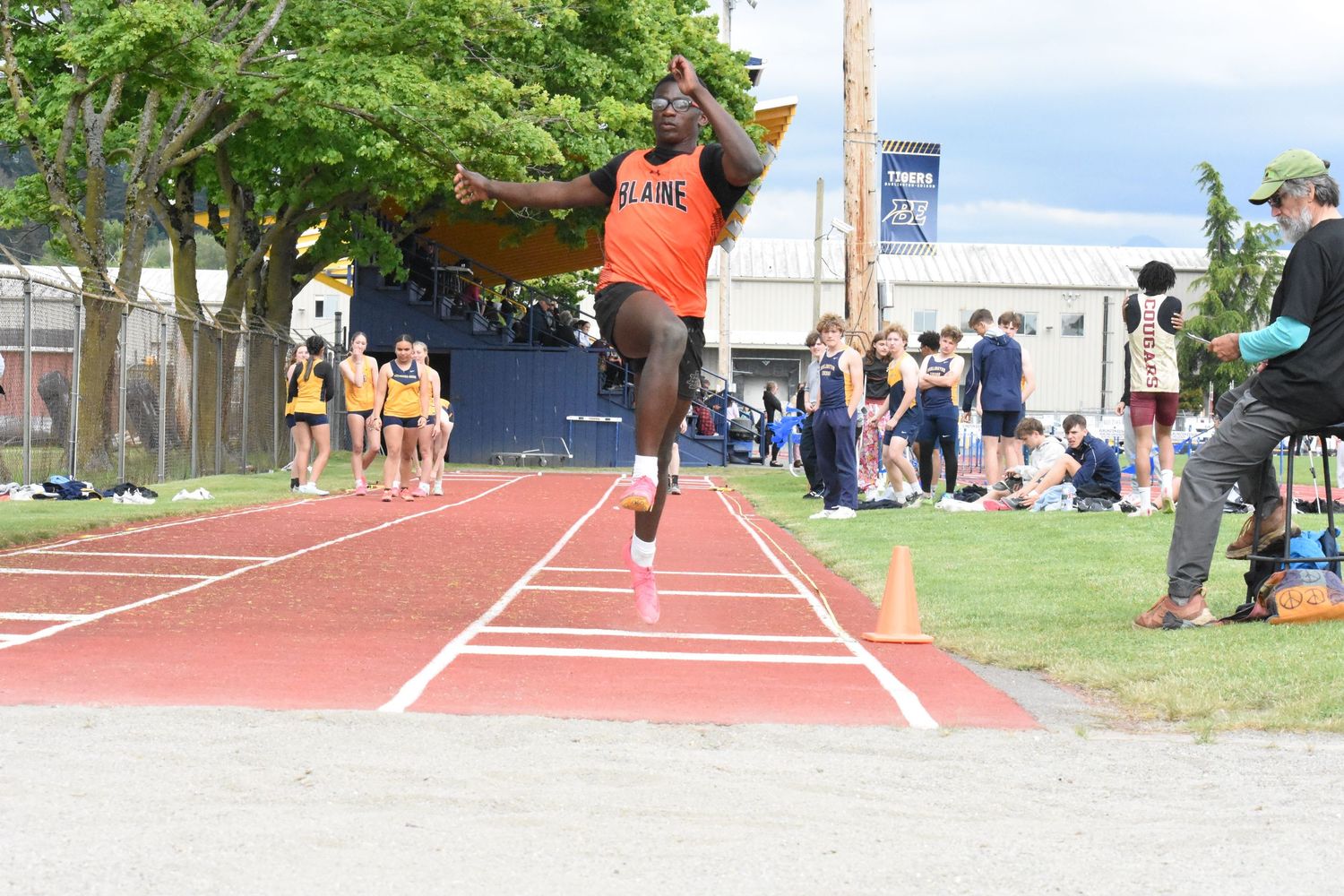 Track and field athlete in orange jersey performs long jump at outdoor competition with spectators watching.