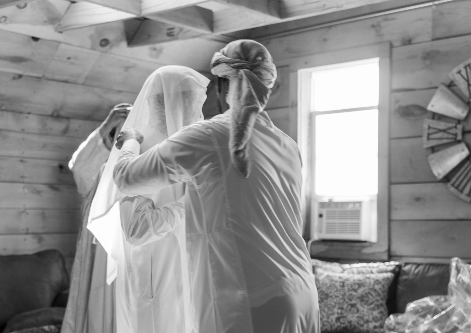 A bride in a white veil being helped by a bridesmaid in a cozy wooden cabin wedding preparation scene.