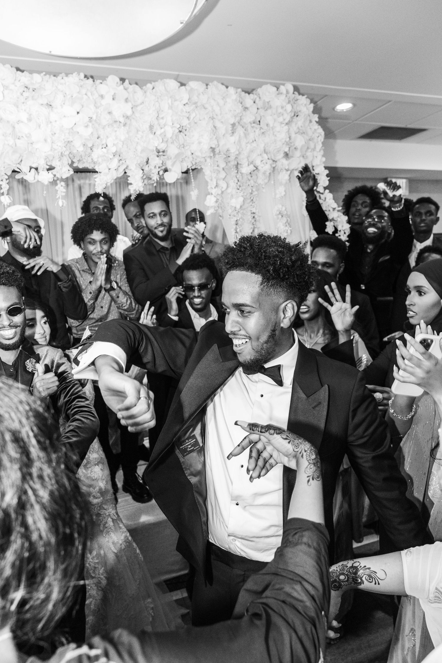 Groom in tuxedo dancing joyfully with wedding guests in black and white celebration photo.