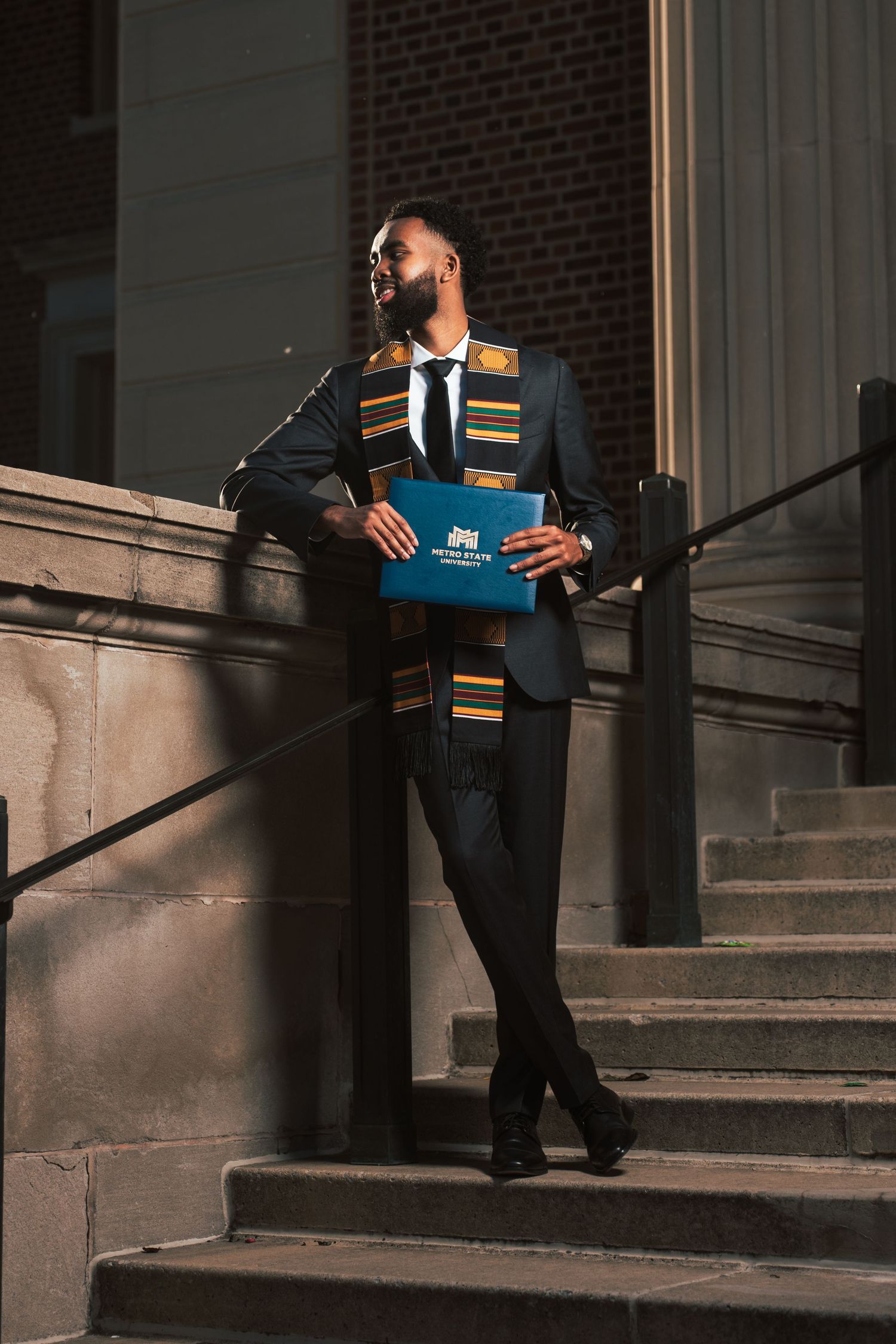 Black male graduate in suit and kente stole holds diploma while posing on building steps outdoors.